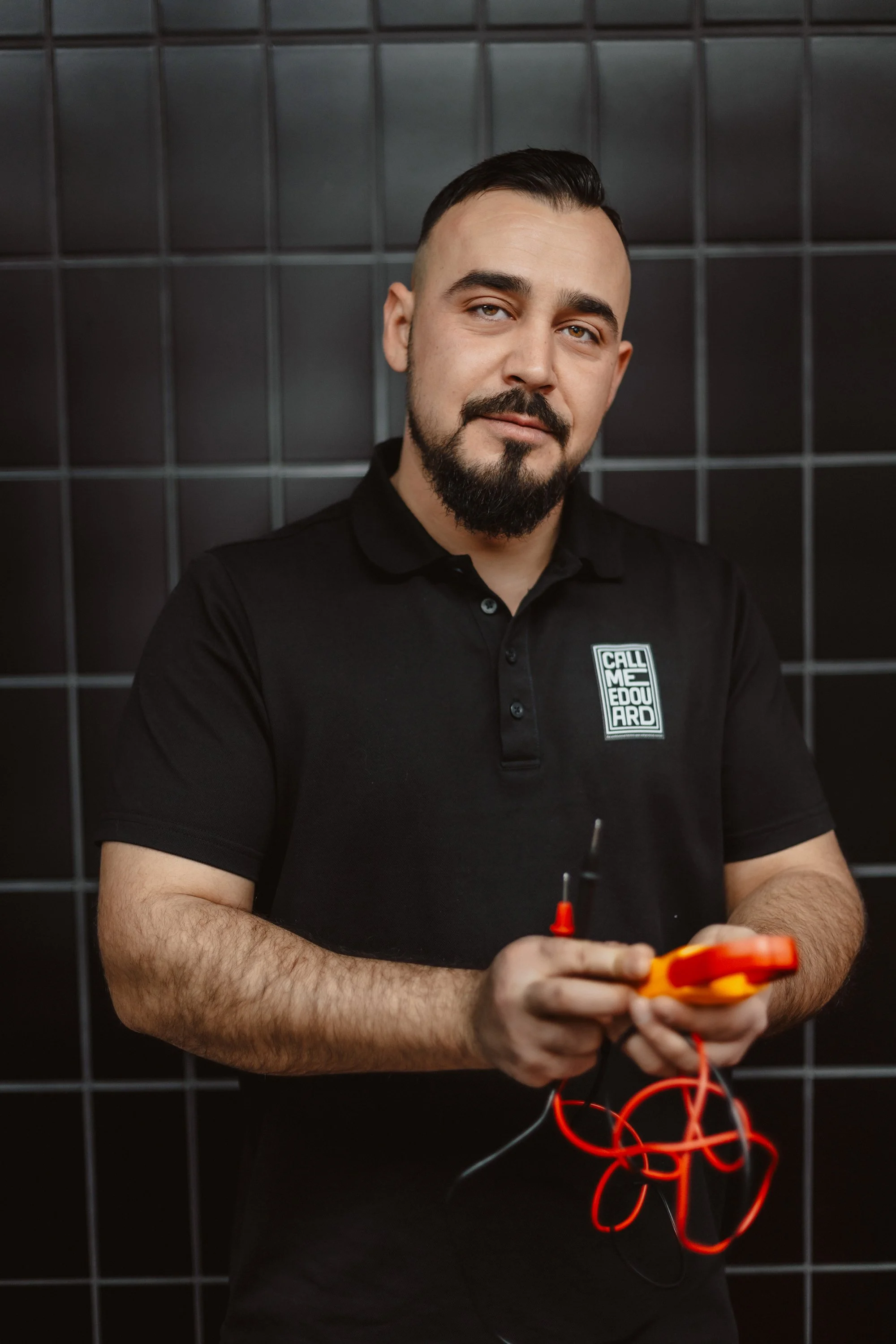 Junger Mann mit dunklem Haar und Bart steht vor einer schwarzen Wand, trägt ein schwarzes Polo mit einem Logo, hält ein elektrisches Messgerät in den Händen.