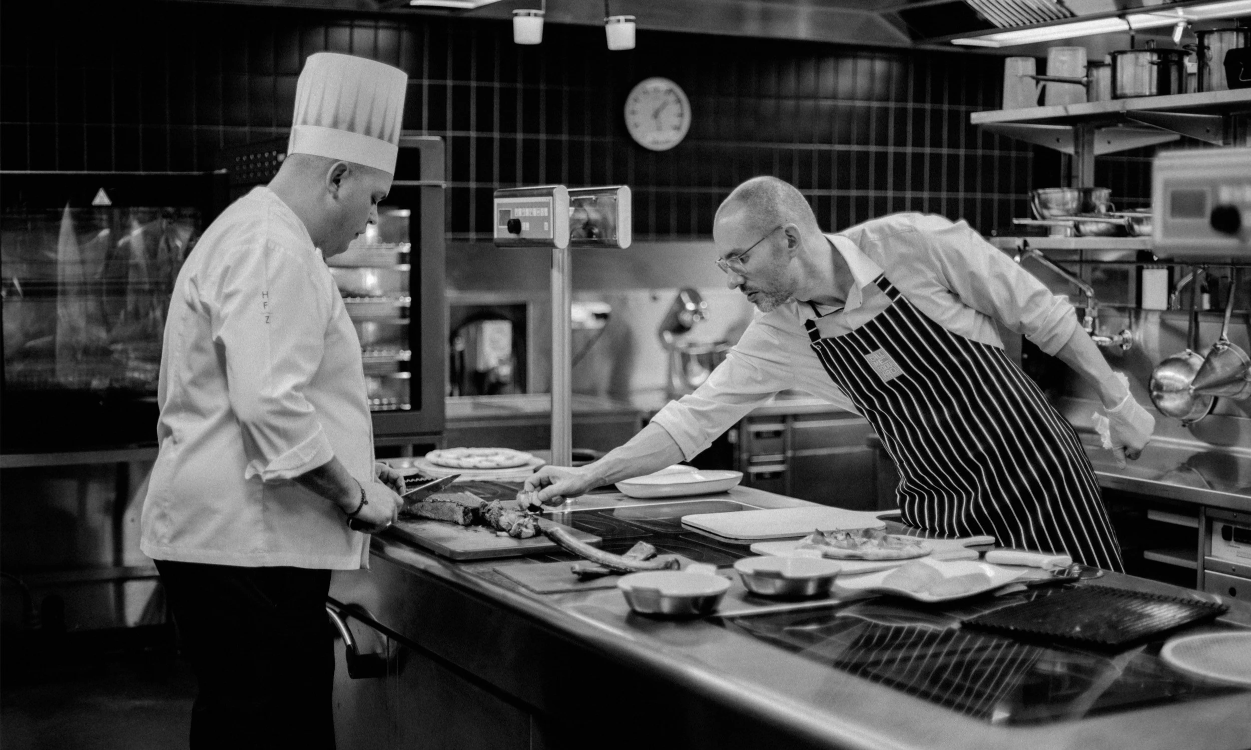 Ein Koch in einer Küche zeigt einem anderen Koch einen Fleischschnitt auf einem Schneidebrett, im Vordergrund. Beide tragen Kochkleidung, einer trägt eine weiße Mütze, der andere eine Schürze mit Streifen.