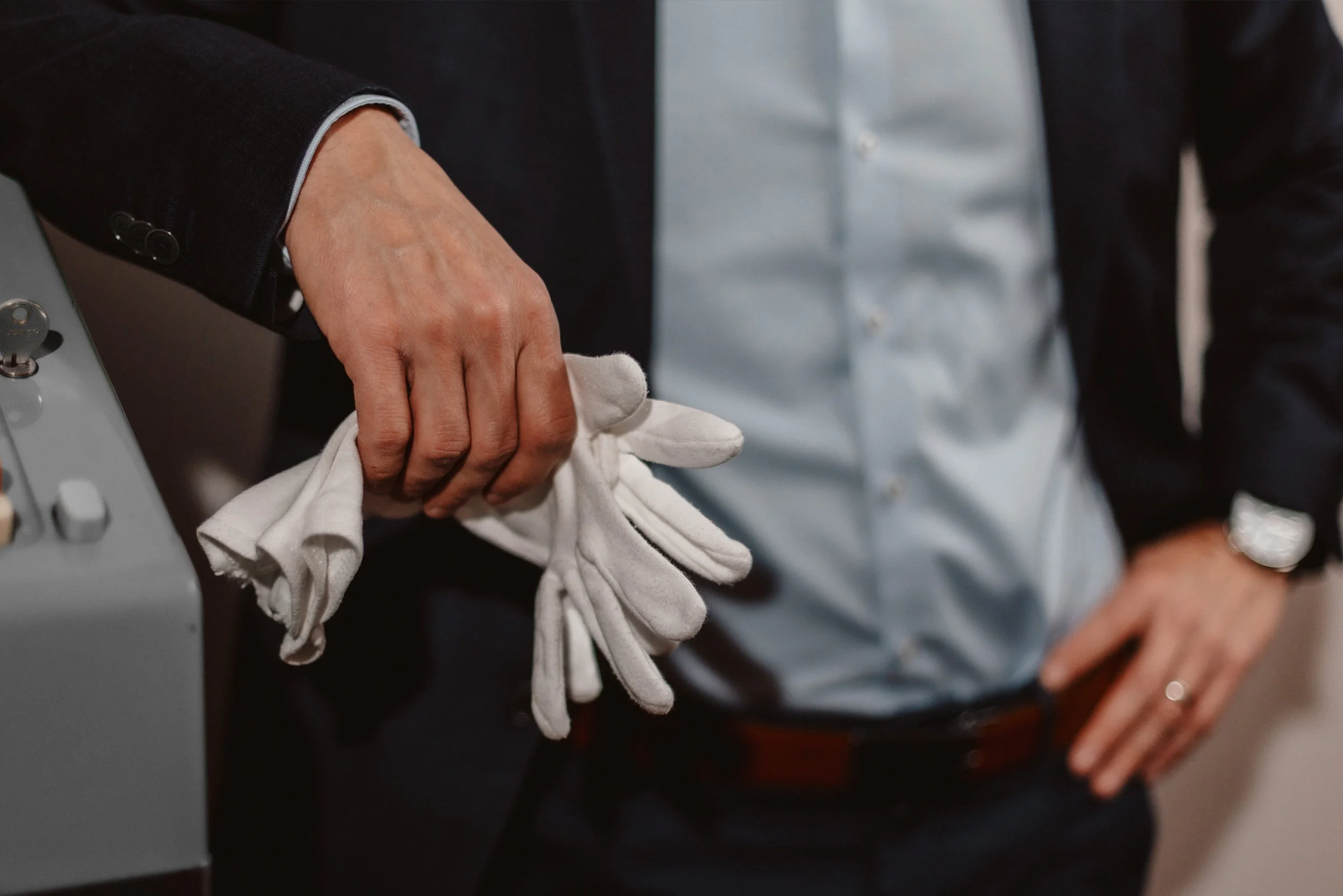 Hand einer Person, die weiße Handschuhe anzieht, vor einem grauen Gerät, mit einer Person im schwarzen Anzug und weißem Hemd im Hintergrund.