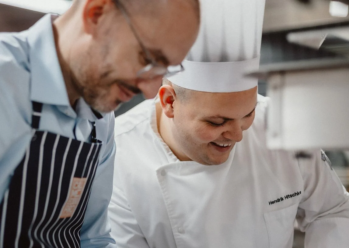 Zwei Köche, einer in weißem Kochoutfit und Kochmütze mit Namen Hendrik Hitsche, der andere in Schürze und Brille, arbeiten in einer modernen Küche.