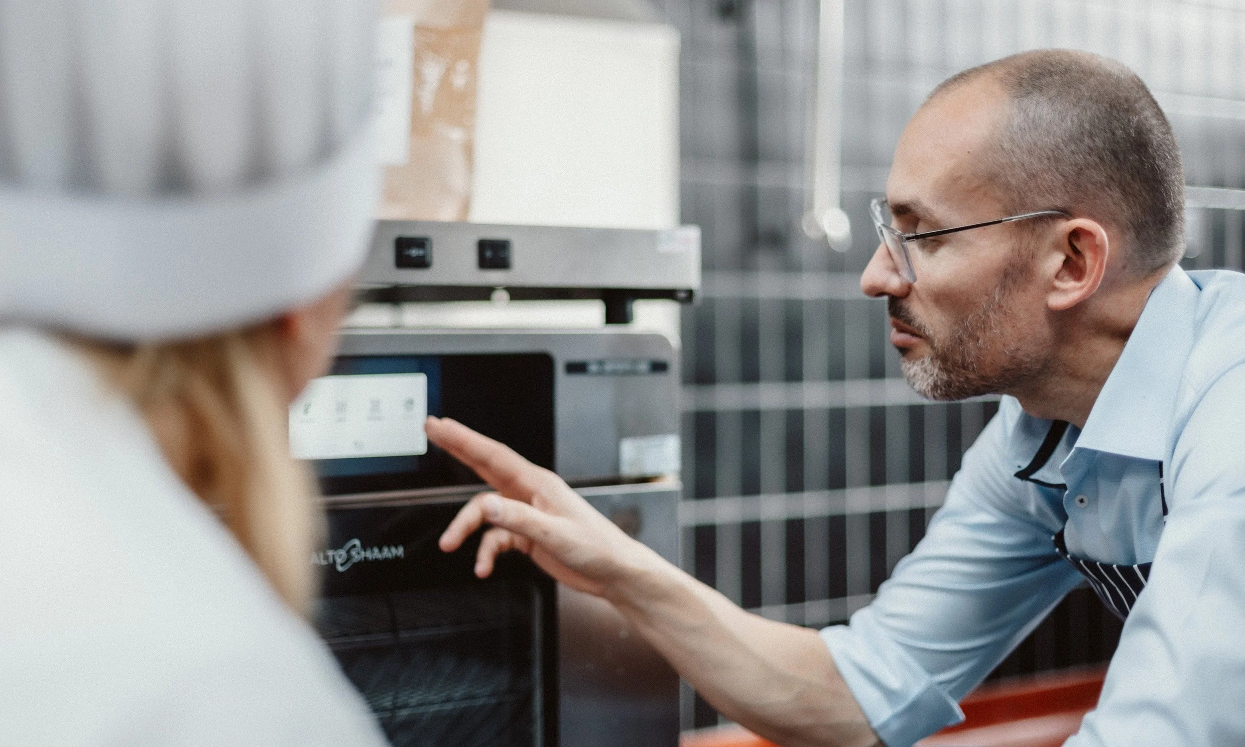 Ein Mann überprüft die Einstellungen eines Ofens in einer professionellen Küche, während eine Frau mit Kochmütze zuschaut.