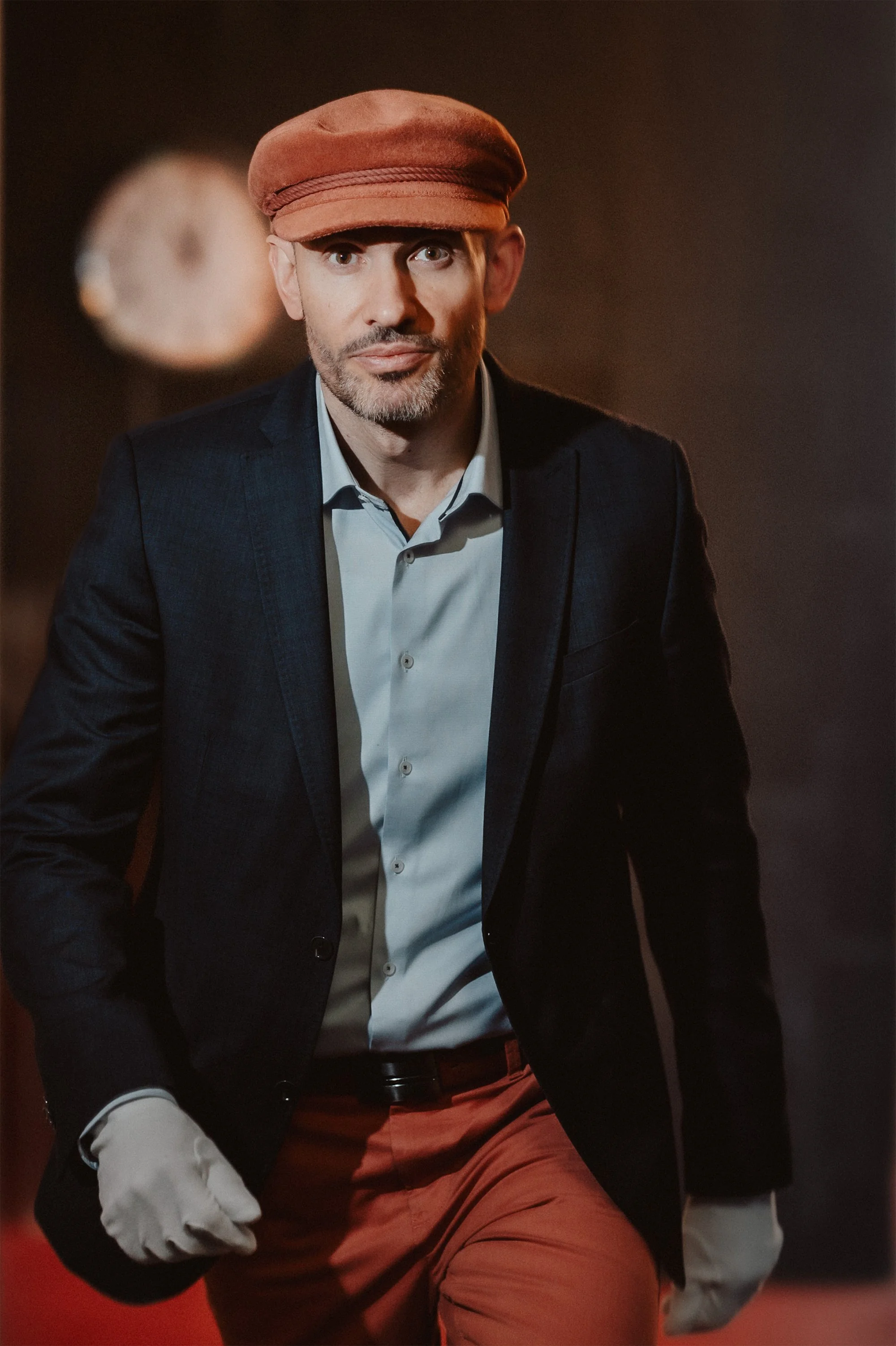 Ein Mann in formeller Kleidung mit schwarzem Anzug, hellblauem Hemd, braunen Hosen, brauner Baskenmütze, weiße Handschuhe steht vor dunklem Hintergrund.