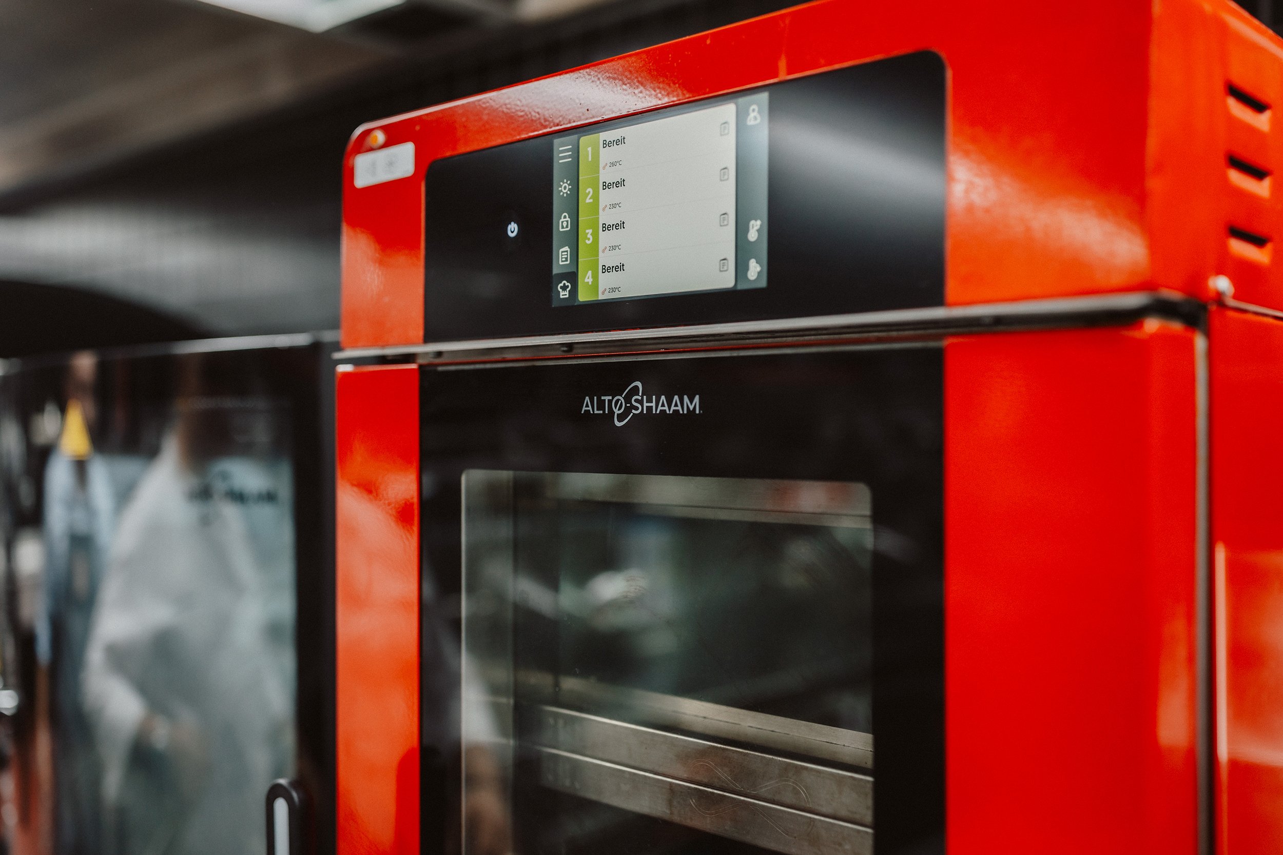 Roter Ofen mit digitalem Steuerungspanel, das verschiedene Betriebszustände anzeigt, im Hintergrund sind Küchenutensilien sichtbar.