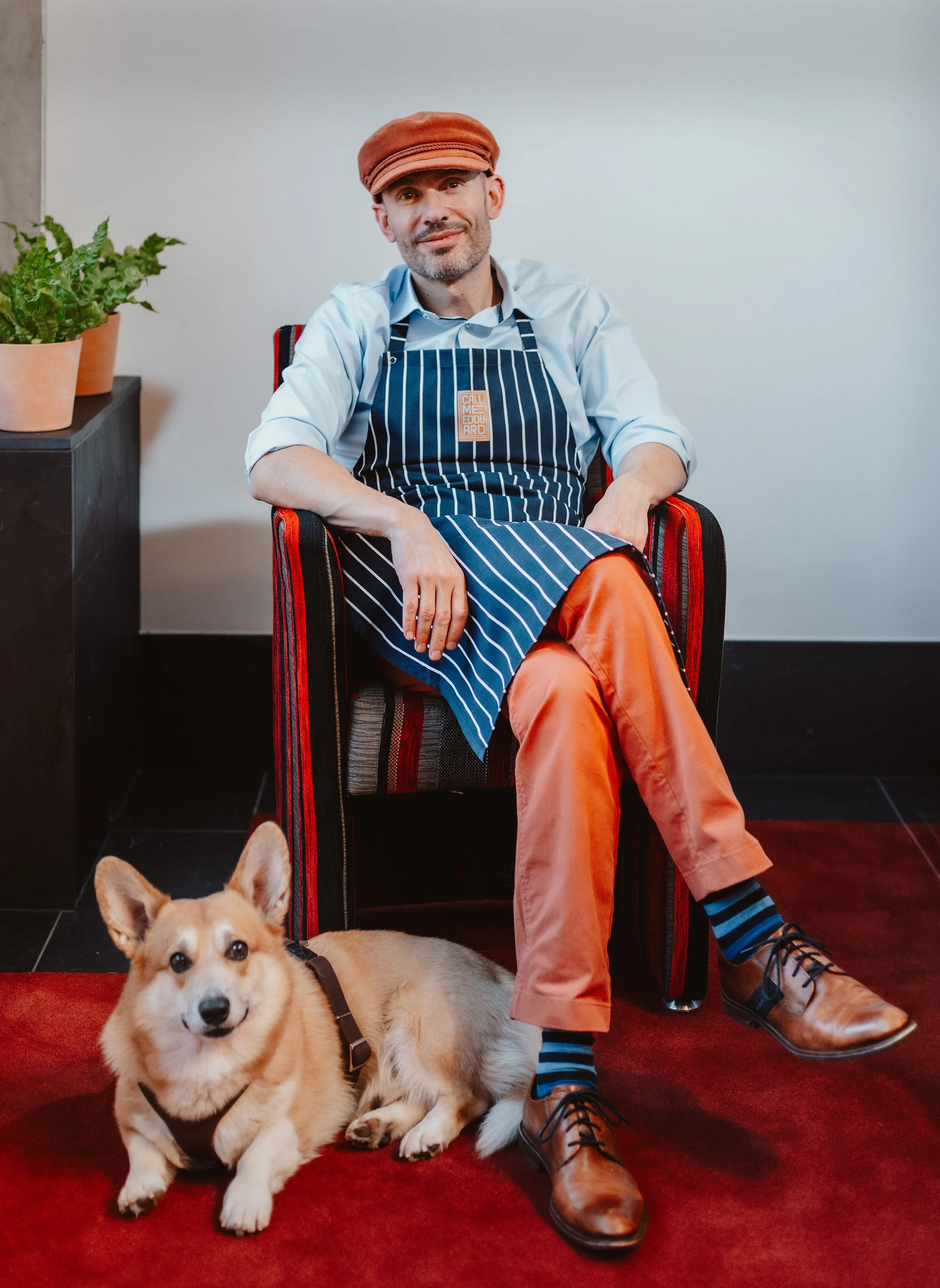 Ein Mann sitzt entspannt in einem roten Stuhl, trägt einen karierten Hut, ein hellblaues Hemd, eine blaue Schürze mit weißen Streifen und orange Hosen. Ein Hund liegt auf dem roten Teppich vor ihm.