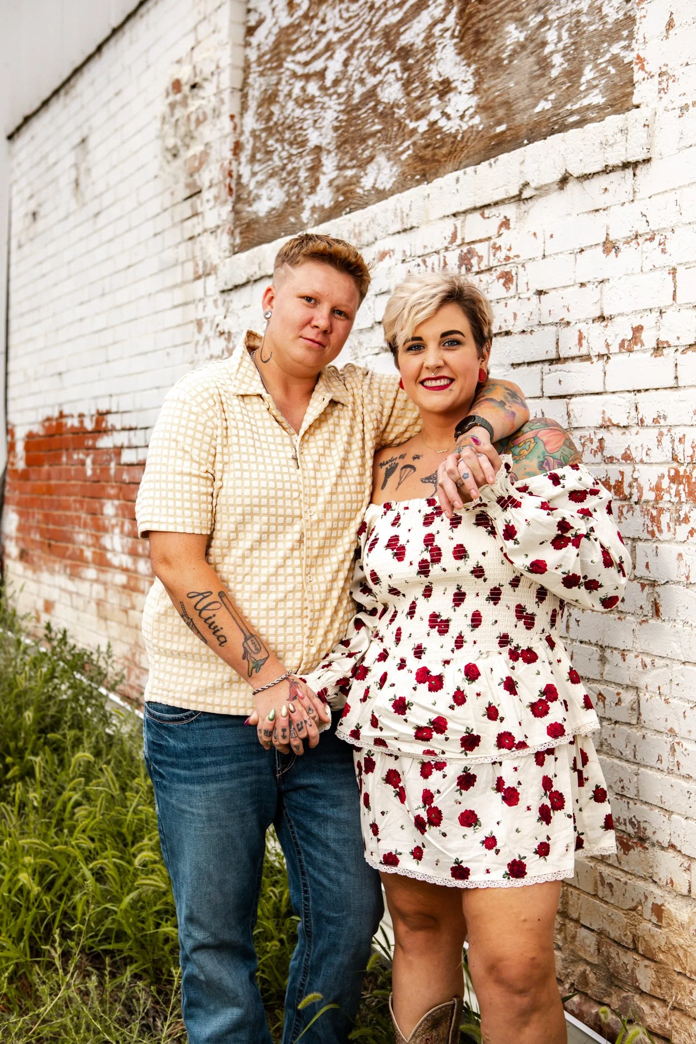 Two women standing close together outdoors against a weathered brick wall, holding hands and smiling, one with short blonde hair and tattoos, wearing a floral off-shoulder dress, and the other with short hair, wearing a short-sleeved checkered shirt 