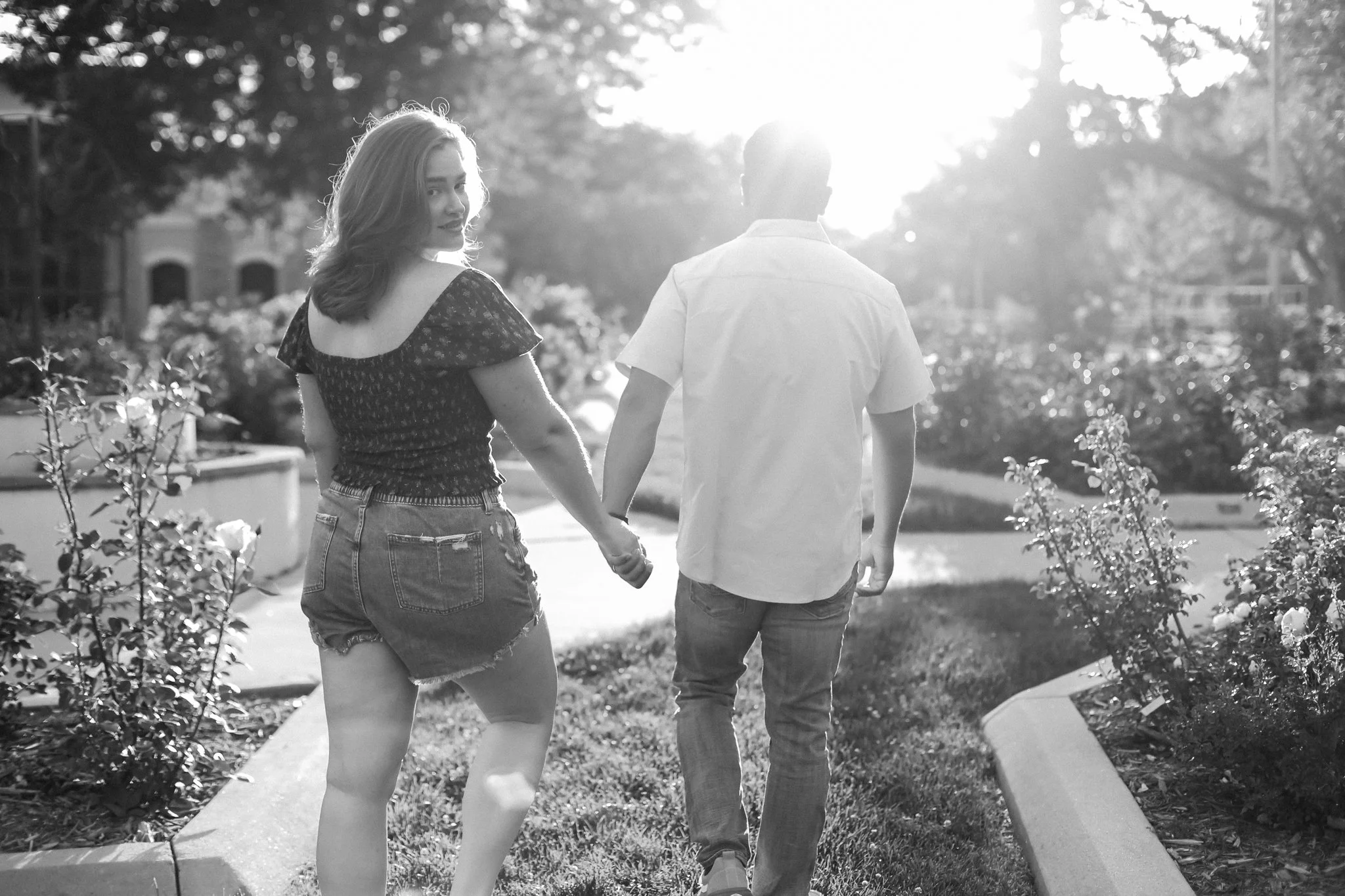 A couple holding hands and walking in a park during sunset, with trees and flowers in the background.