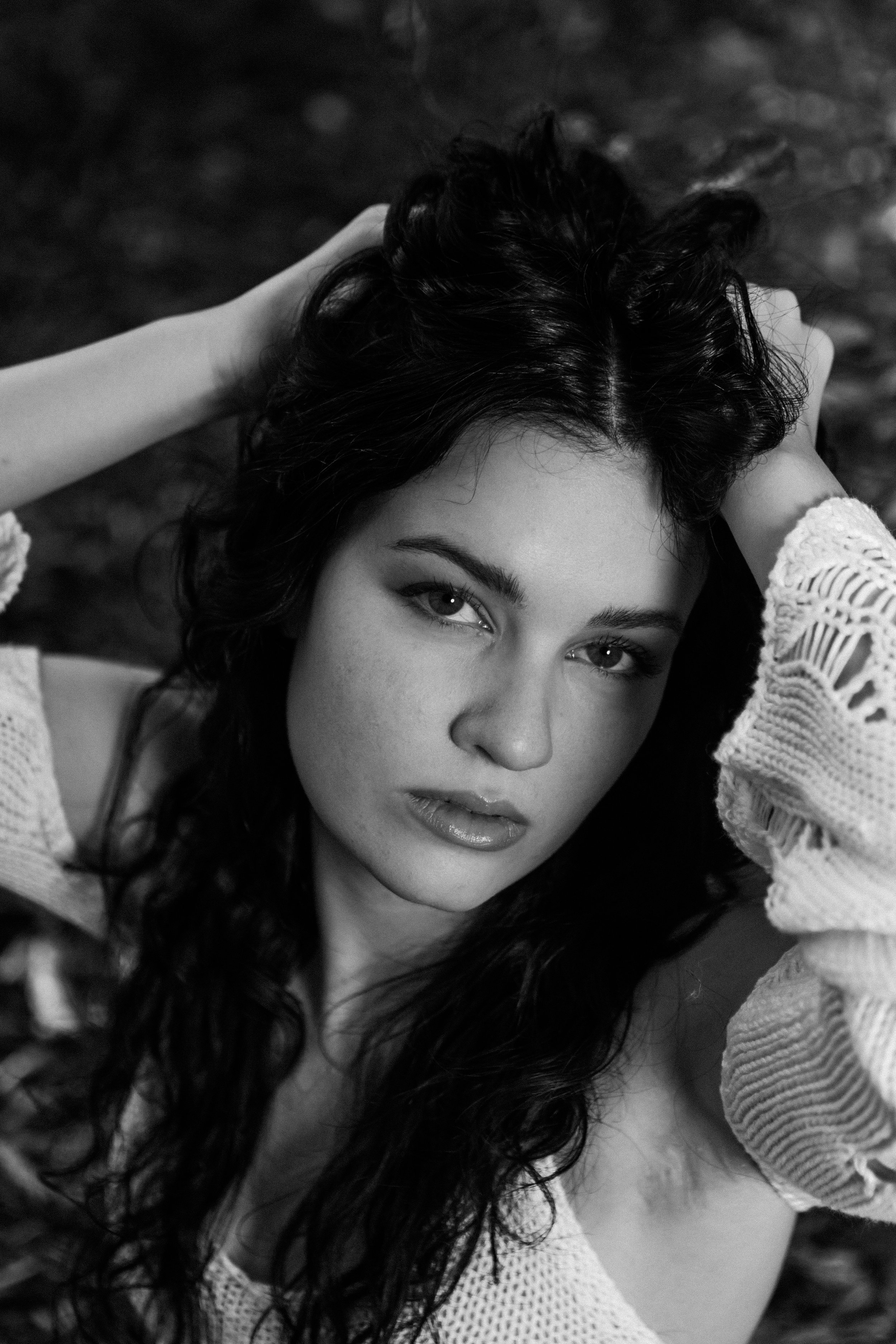 A black and white close-up portrait of a young woman with long, curly hair, looking directly at the camera with her arms raised, hands resting on her head, wearing a light-colored knitted sweater.