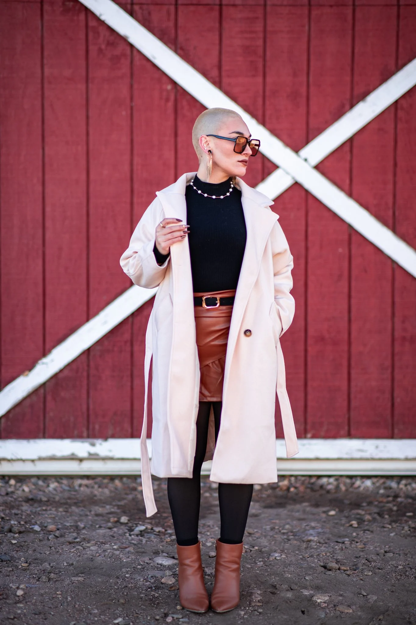 A woman with a shaved head wearing a beige trench coat, black turtleneck, brown leather skirt, black tights, brown ankle boots, large sunglasses, pearl necklace, and earrings stands in front of a red barn with white diagonal stripes.