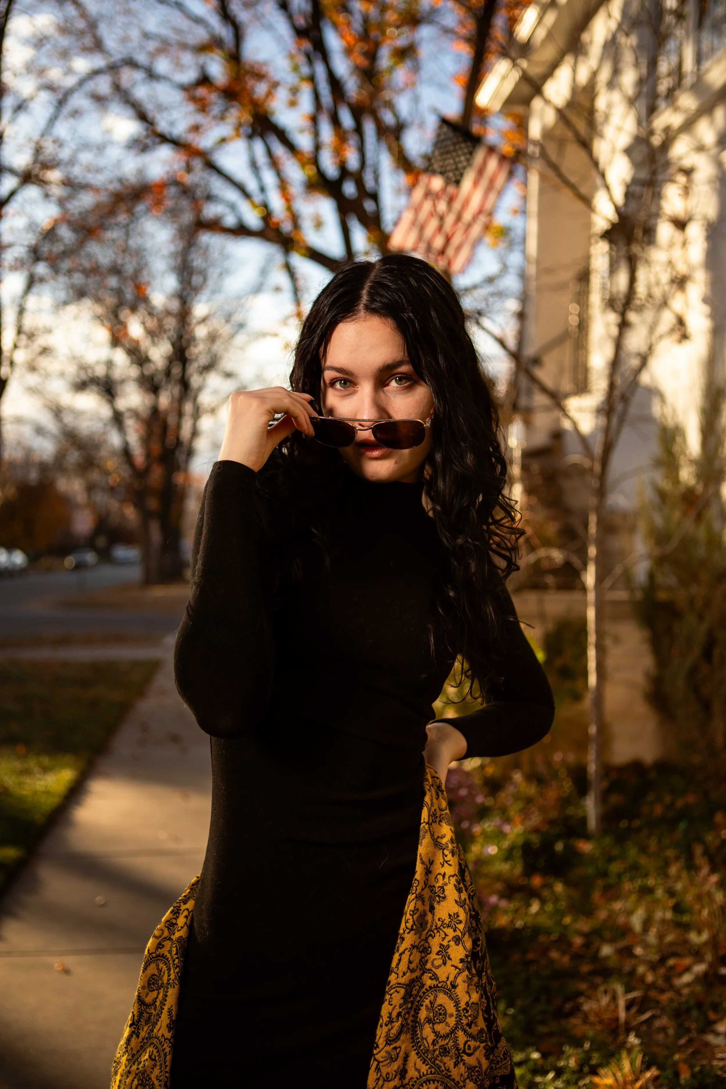 A young woman with black hair and light skin adjusting brown sunglasses on her face outdoors during autumn, standing on a sidewalk with trees and American flags in the background.