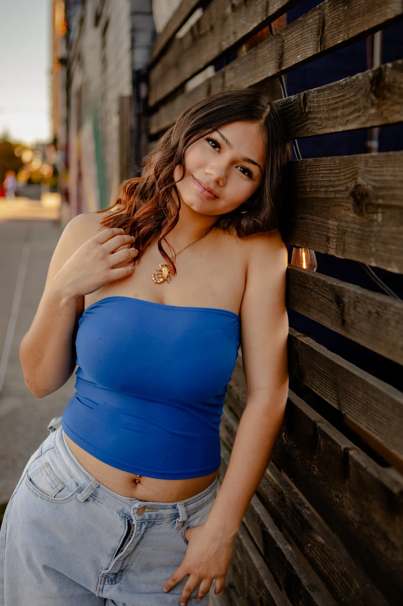 A young woman with wavy brown hair, wearing a blue strapless top and light denim jeans, leaning against a wooden fence during sunset.