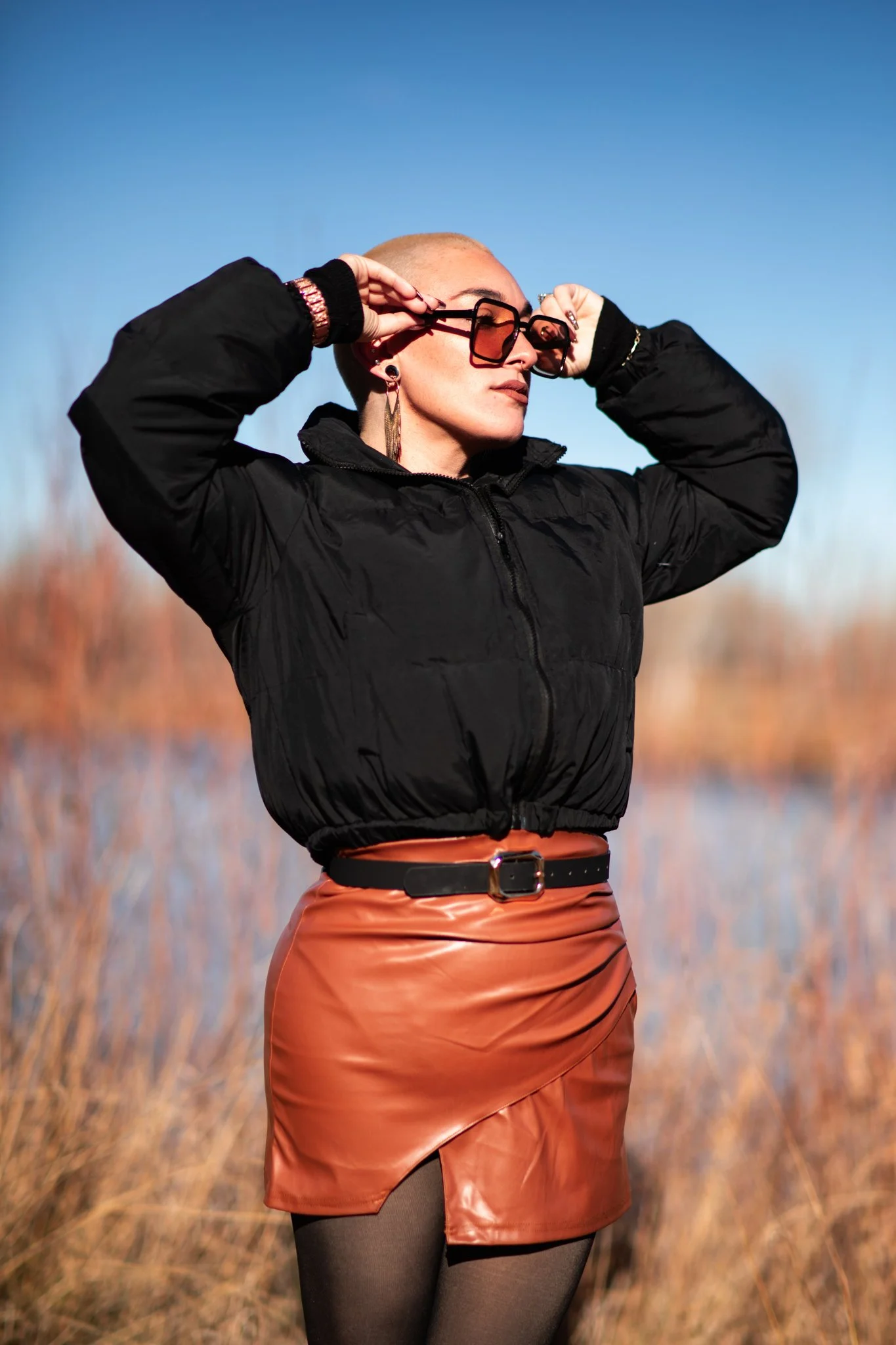A woman with a shaved head and sunglasses adjusting her glasses outdoors during daytime, wearing a black jacket, a tan skirt, and tights, with a blurred natural background.