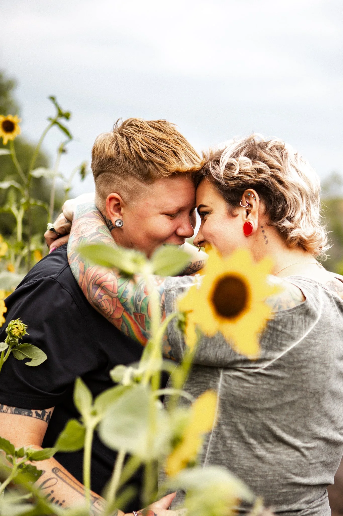 Two women with tattoos and piercings embracing in a sunflower field, noses touching, smiling.