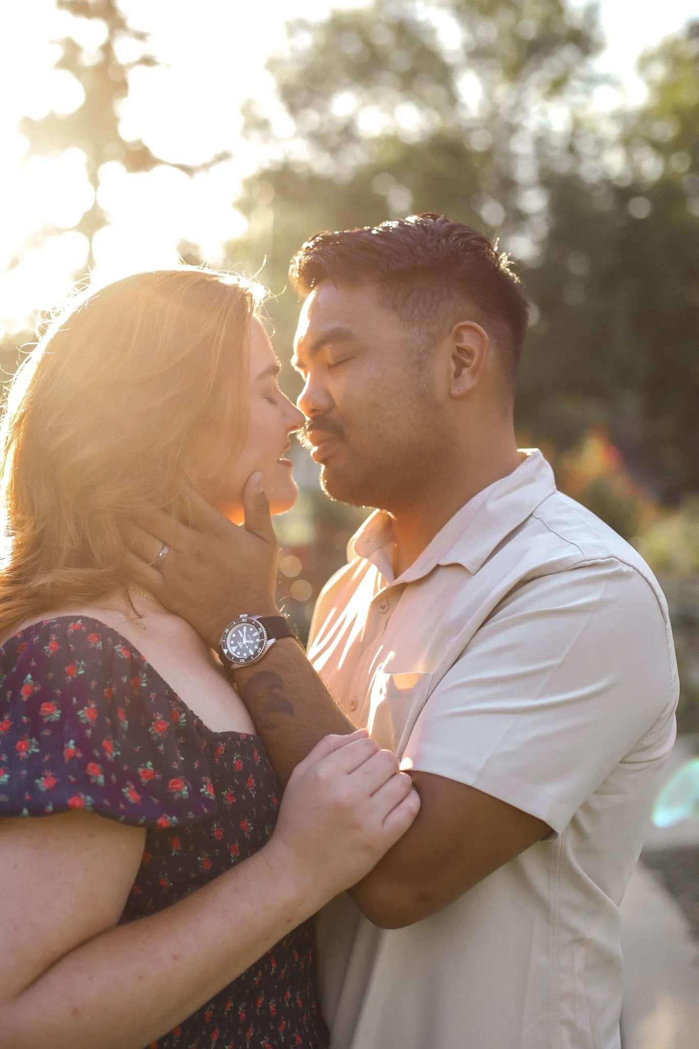 A couple standing close together outdoors during sunset, with the woman gently holding the man's face as they look into each other's eyes.