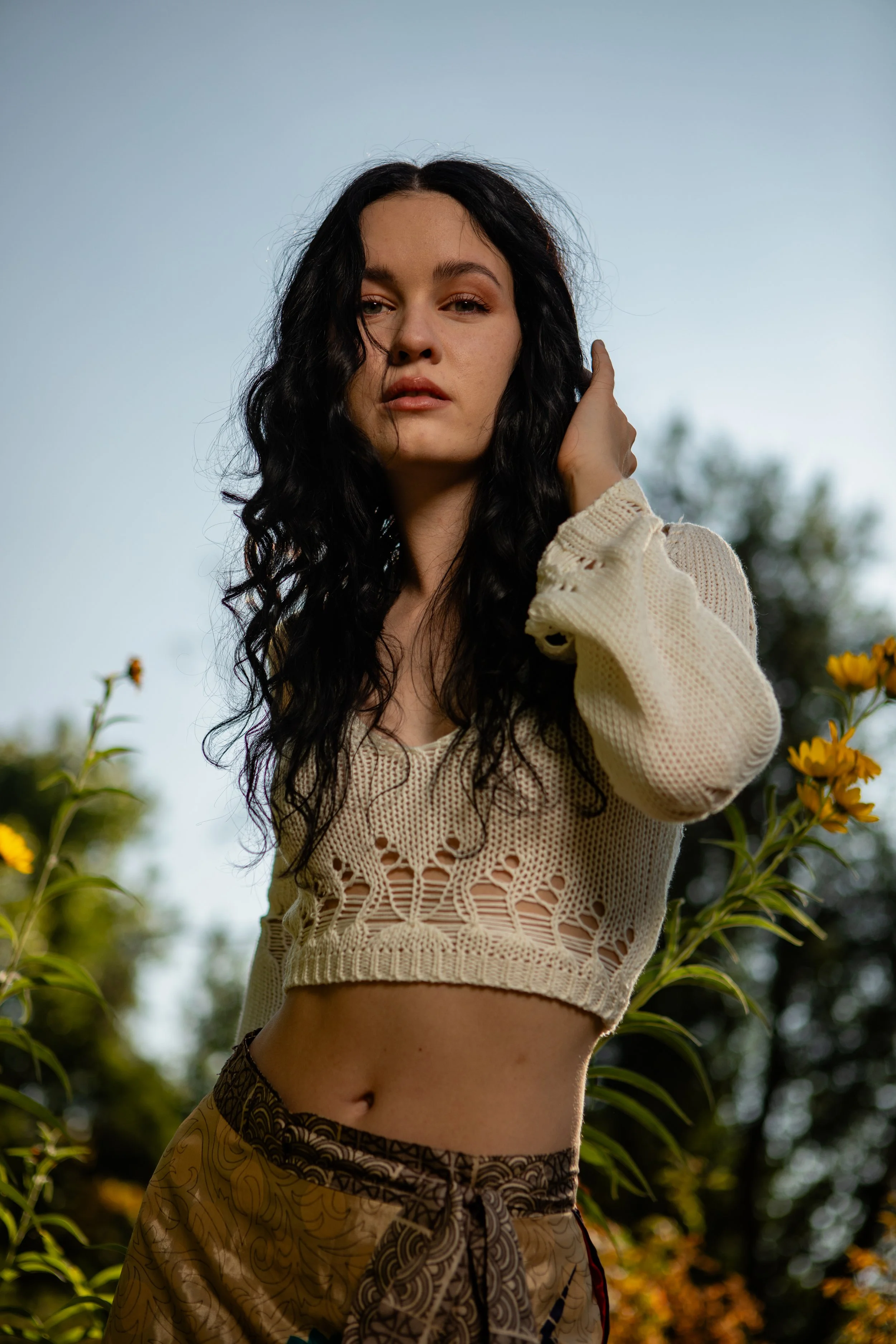 A young woman with black curly hair posing outdoors against a background of yellow flowers and green foliage during daytime, wearing a cream-colored crochet top and patterned brown pants.