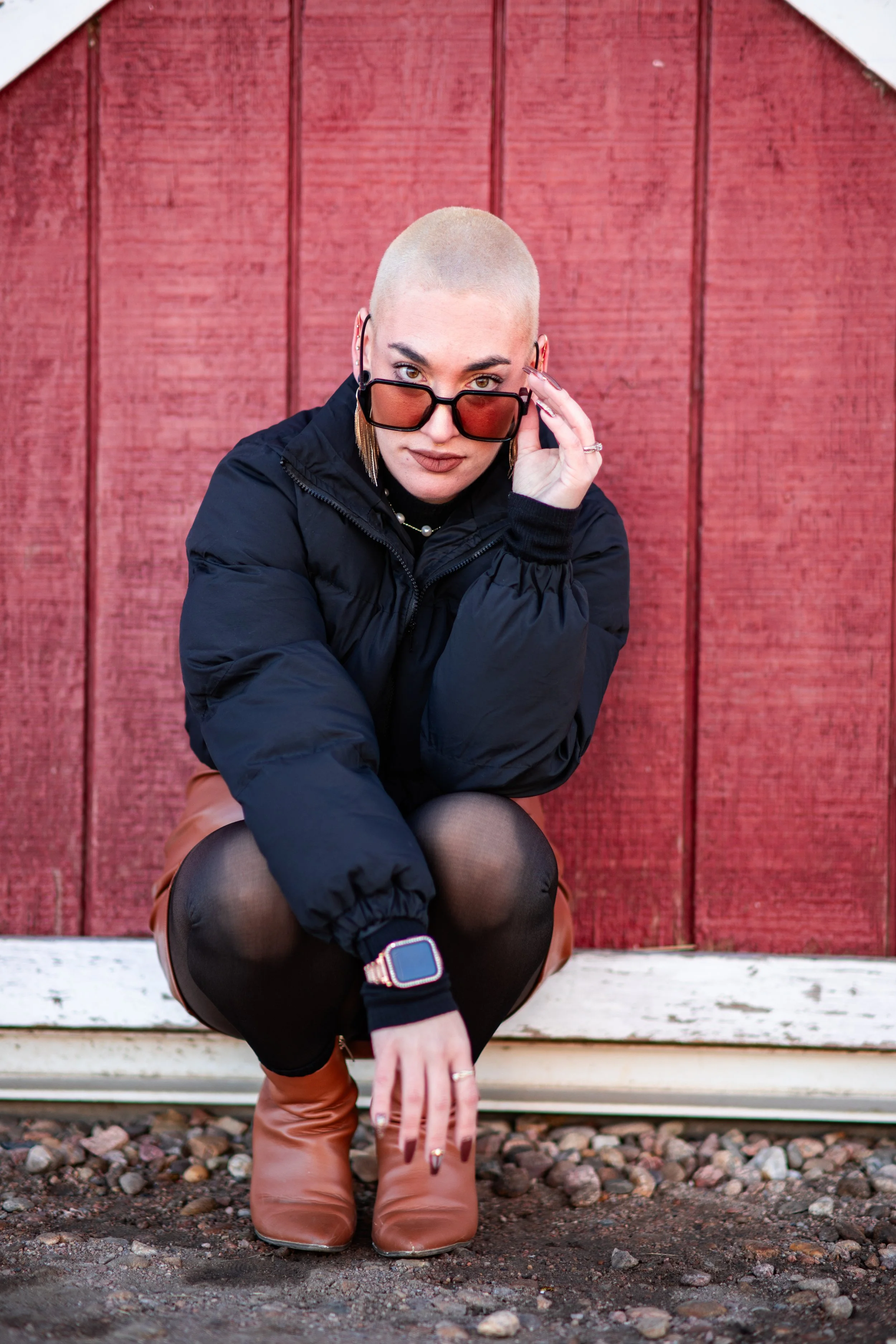 A woman with a buzz cut hairstyle squatting outdoors in front of a red wooden wall, wearing sunglasses, a black jacket, black tights, a brown skirt, and tan boots, with a smartwatch on her wrist.