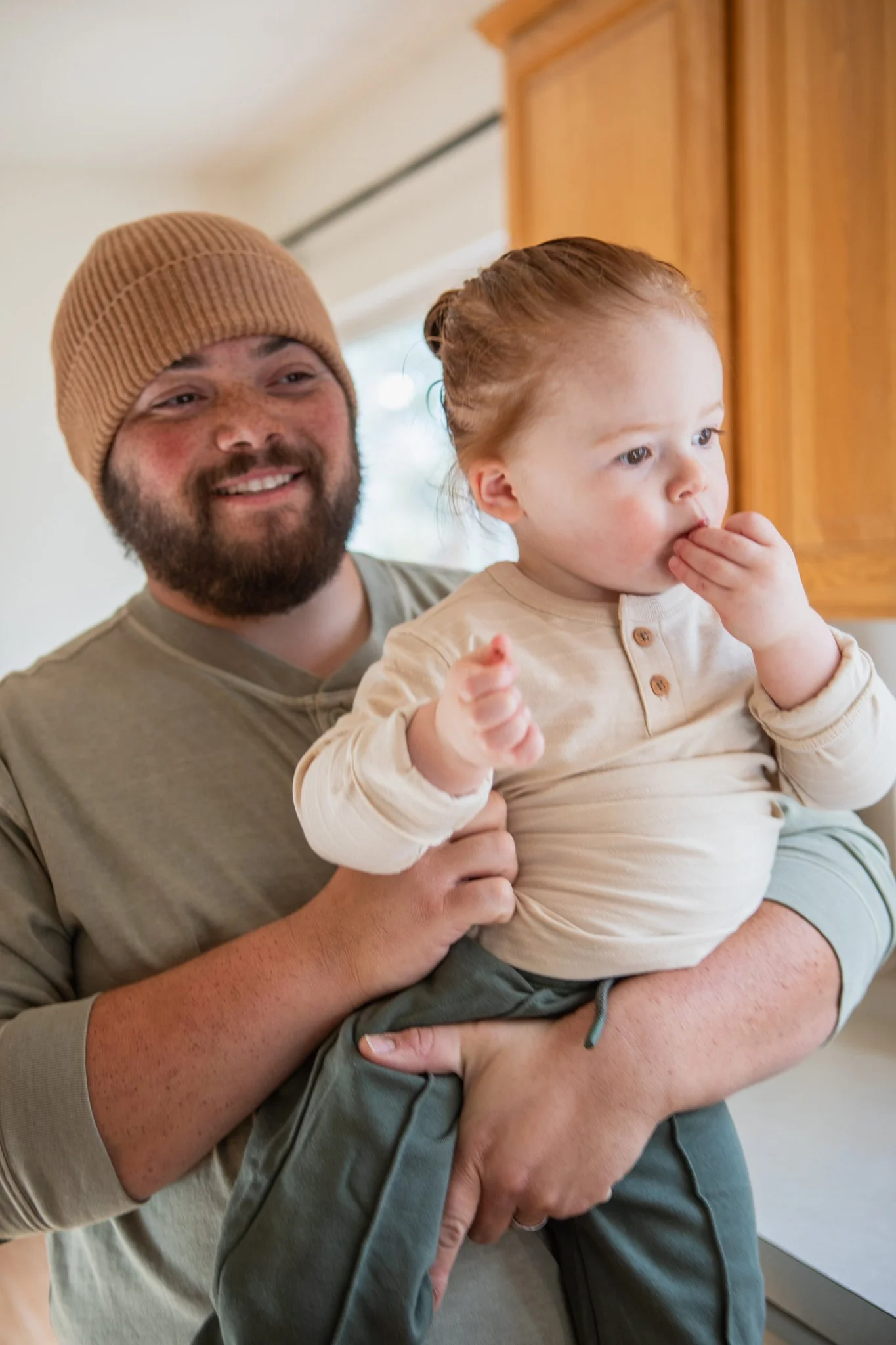 A man with a beard and wearing a brown beanie is holding a young girl with red hair in a kitchen. The man is smiling, and the girl is touching her face with her hand.