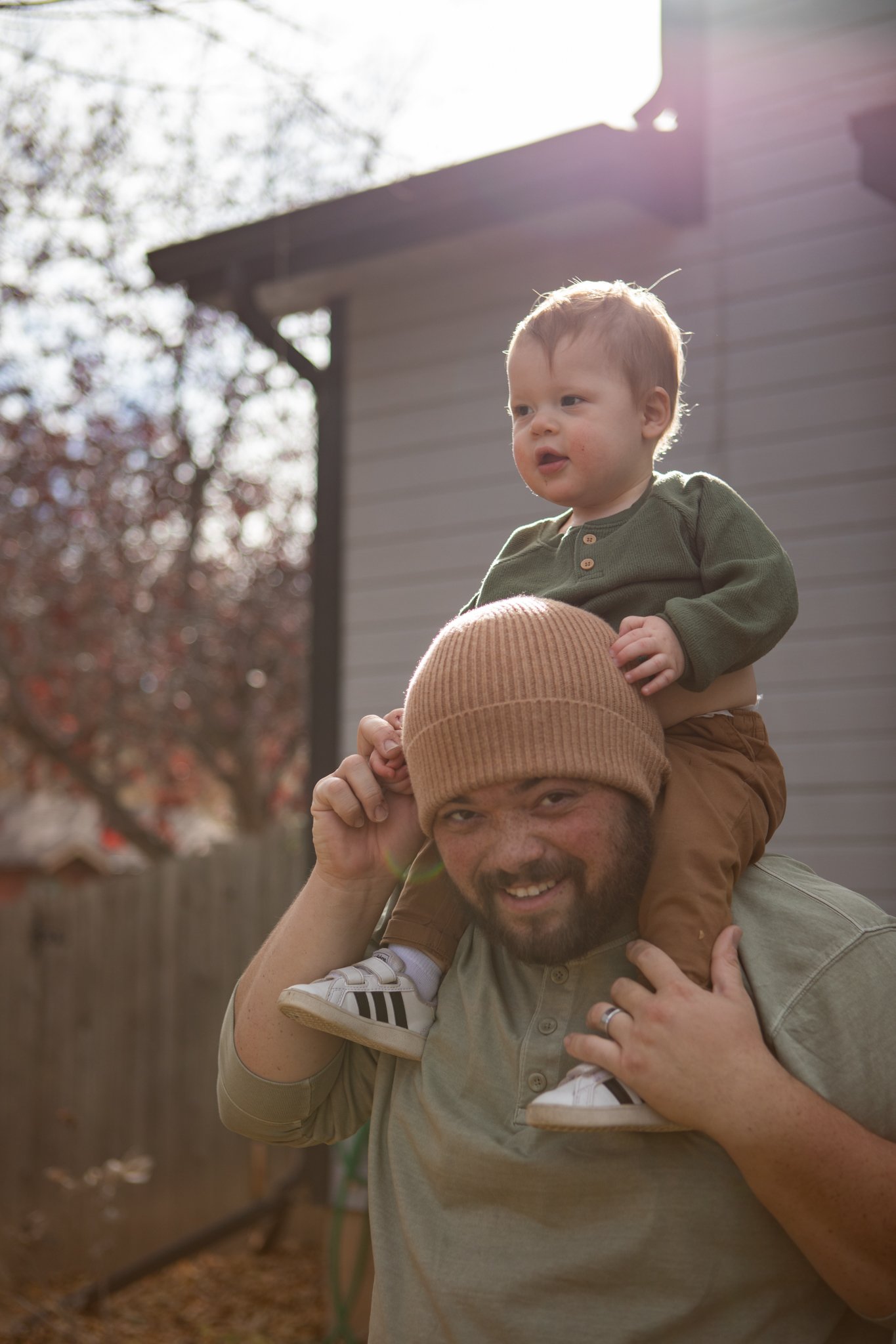 A man with a beard and a brown knit cap carrying a young child on his shoulders in a backyard with a wooden fence and trees in the background, under sunlight.