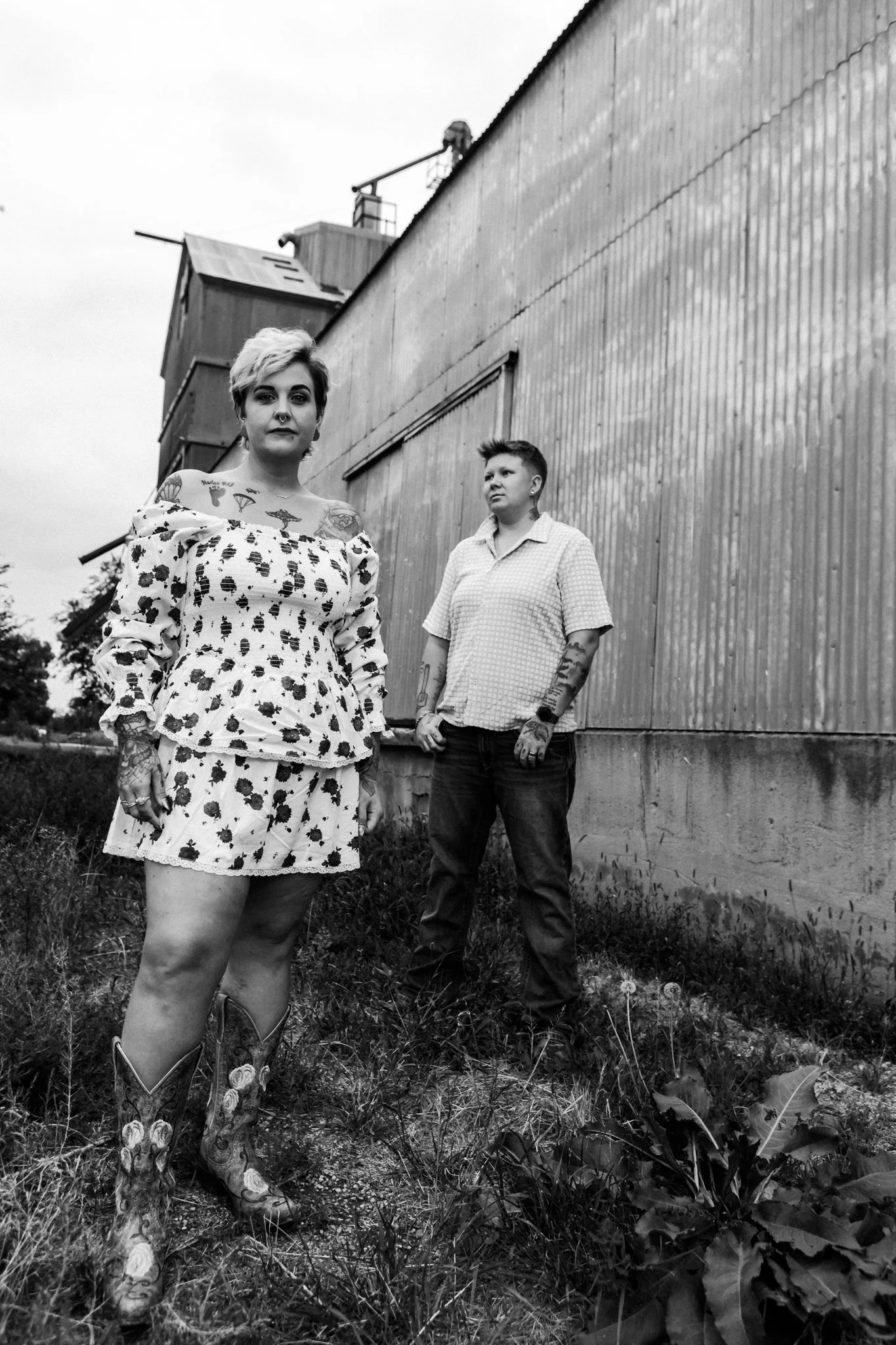 Two women with tattoos standing outdoors near a large wooden building, one facing camera and the other looking away, black and white photo.