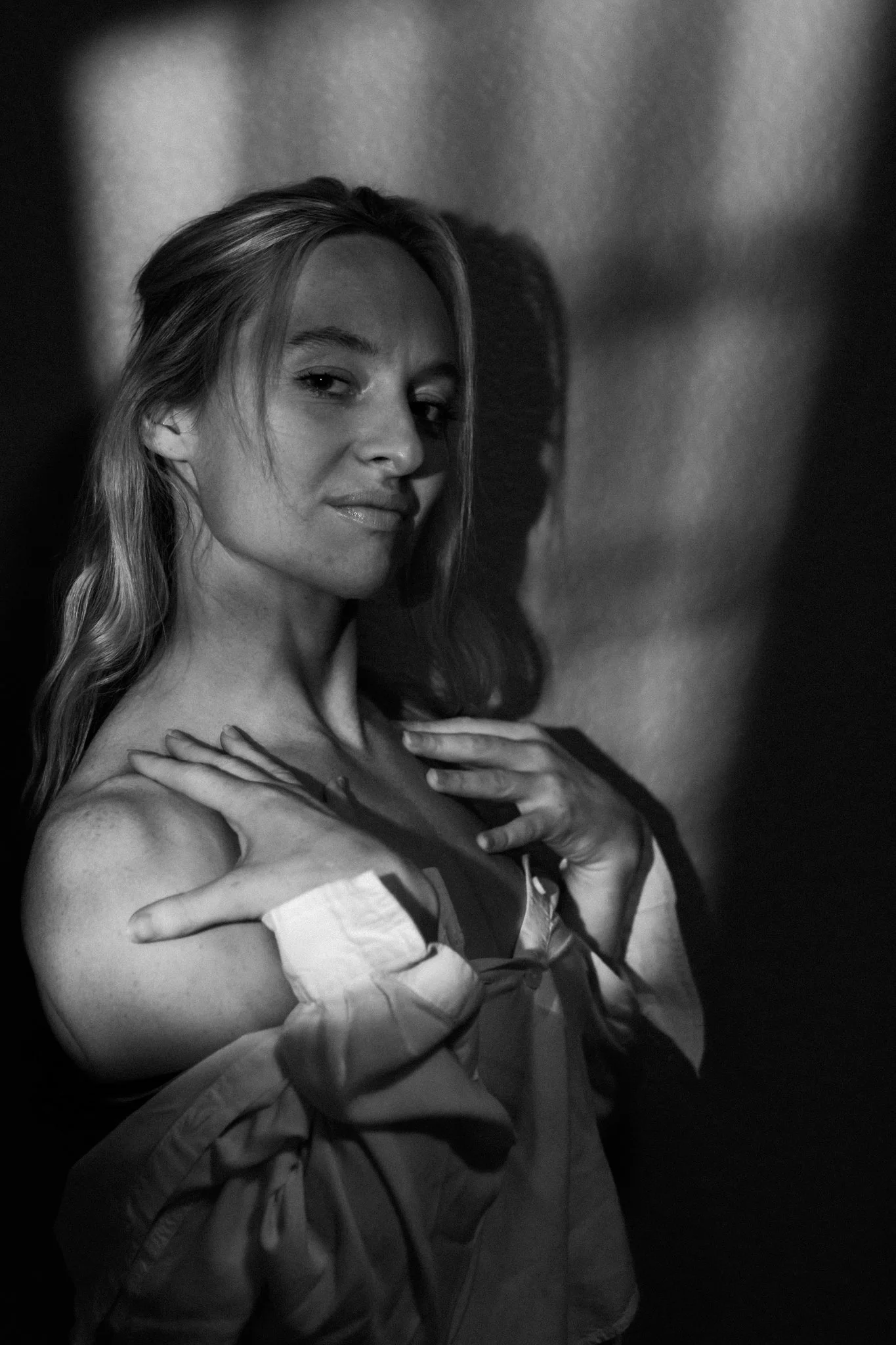 A black and white portrait of a woman with long hair, partially revealing her shoulders and chest, with her hands crossed over her chest and a confident expression.