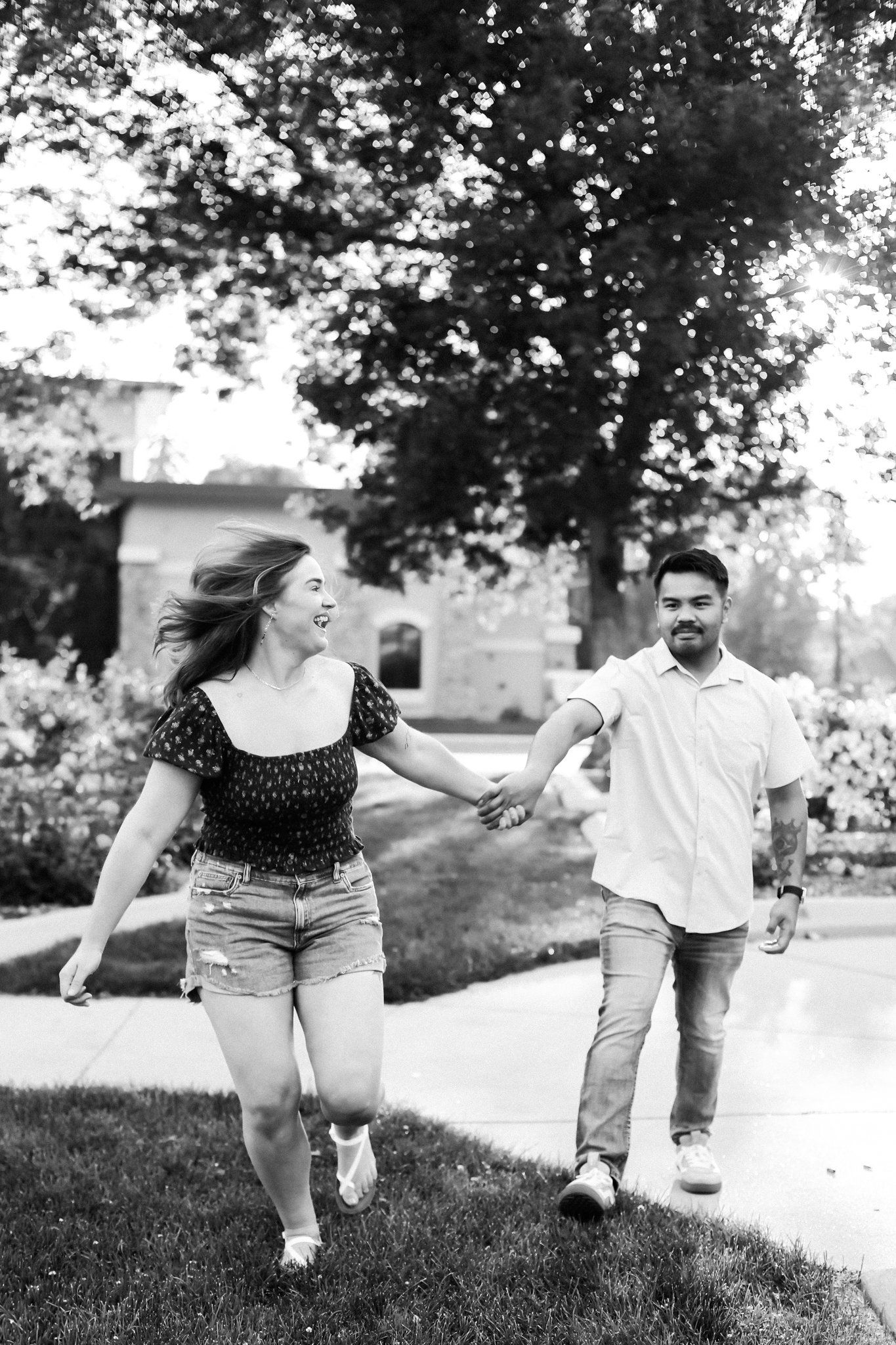 A woman and man holding hands and running outdoors on a sidewalk with trees and houses in the background, black and white photo.