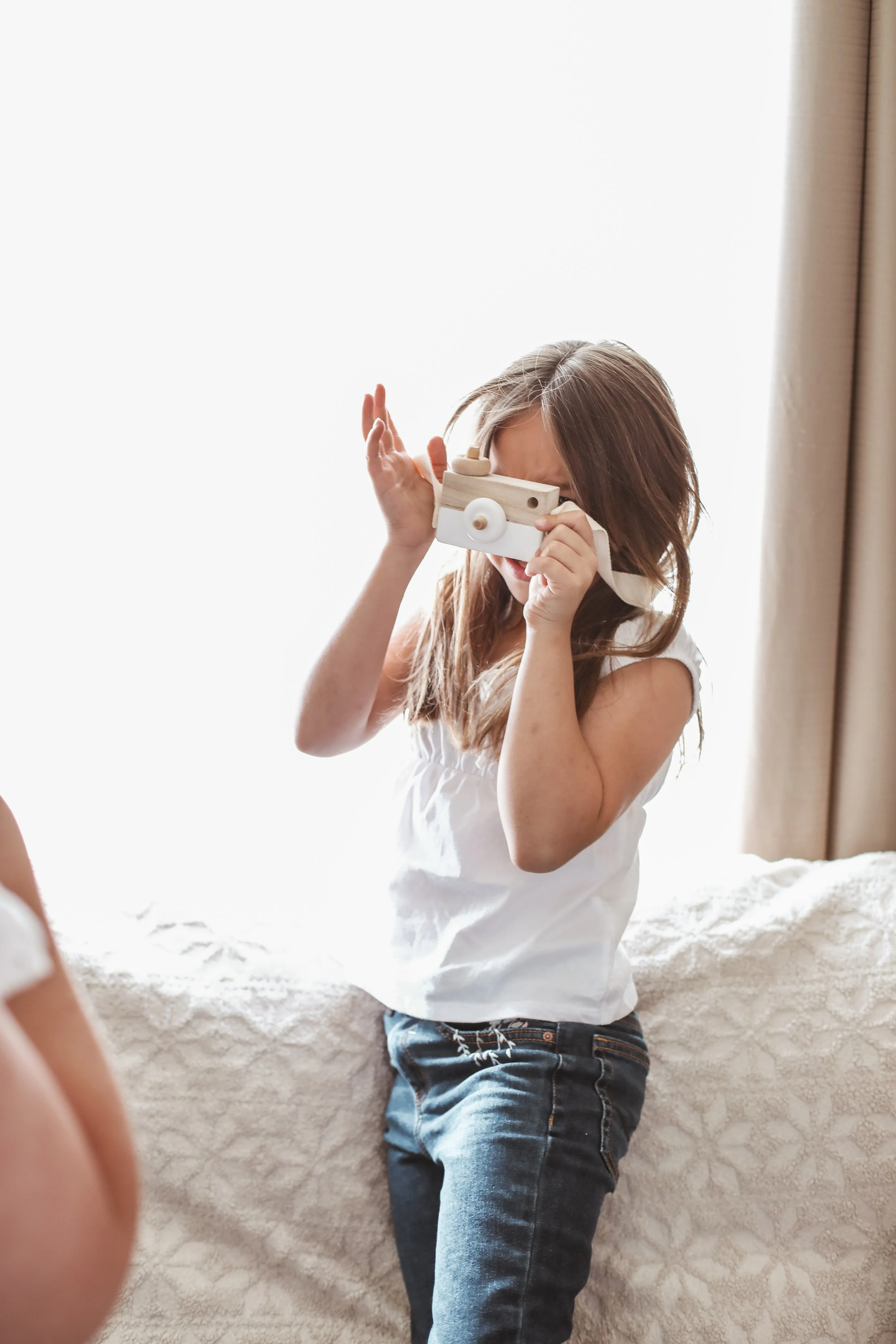A young girl with long brown hair playing with a wooden toy camera near a window with curtains.