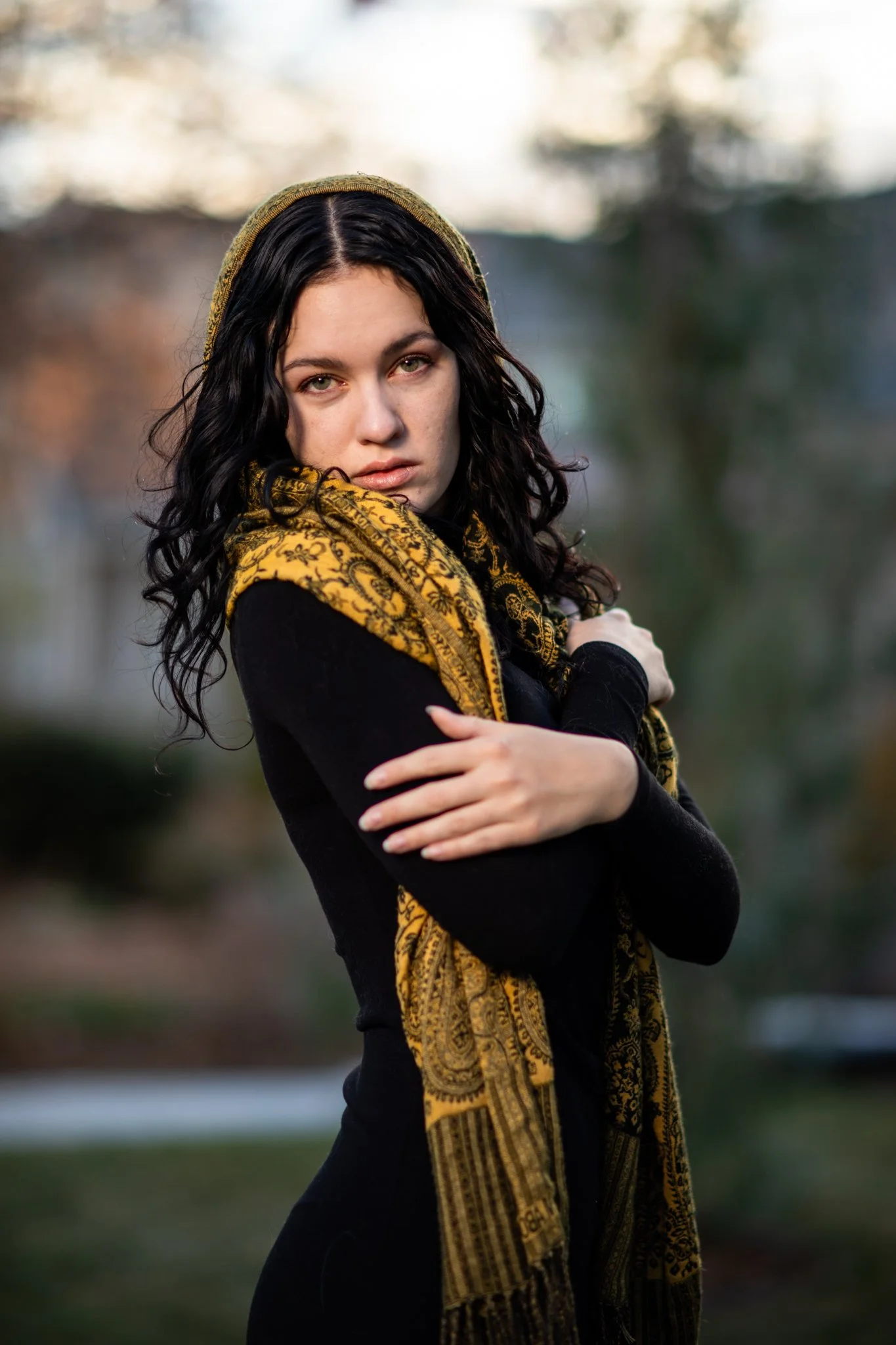 A woman with long dark curly hair wearing a black long-sleeve top and a yellow patterned scarf, standing outdoors with a blurred background.