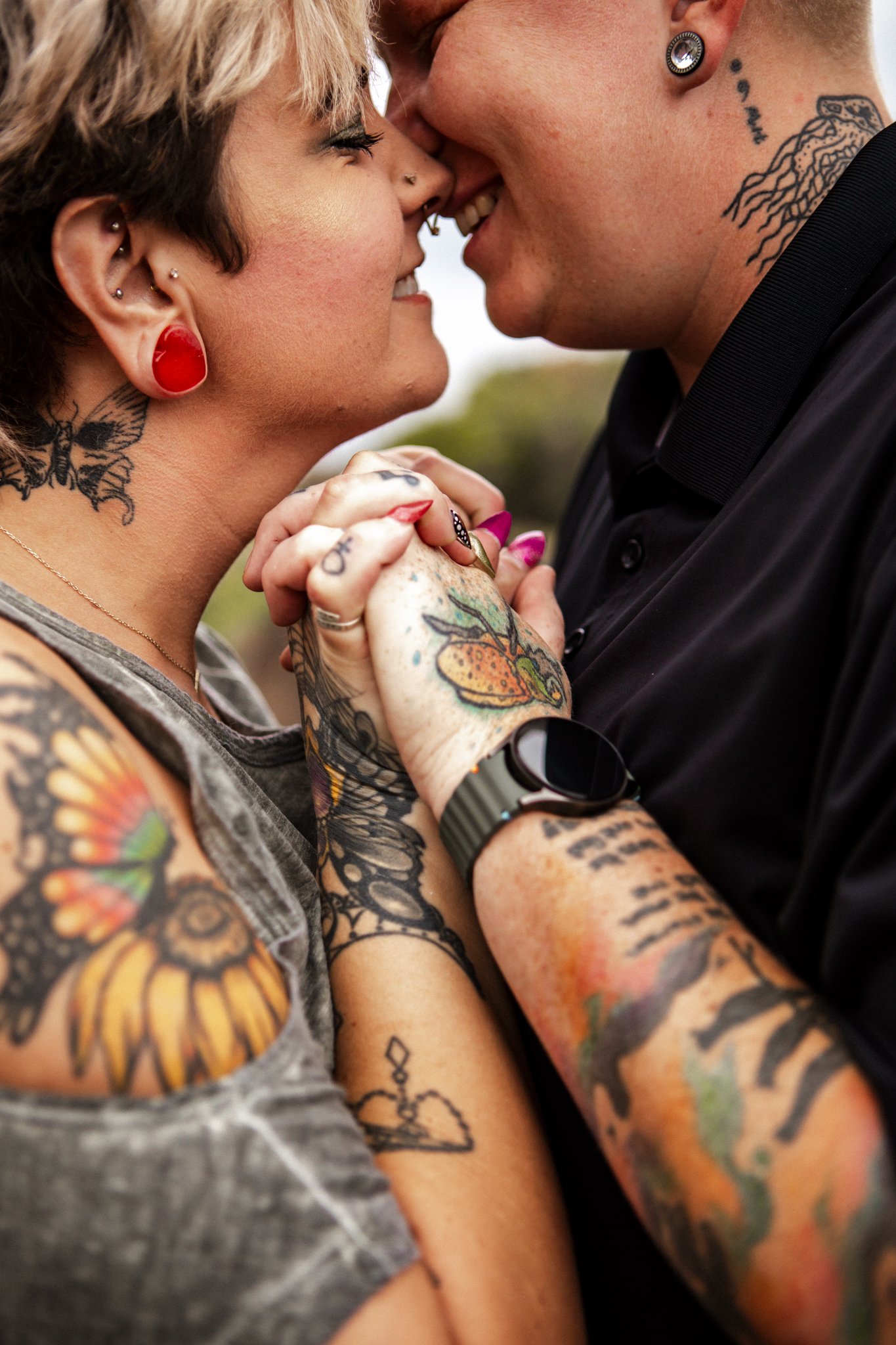 A close-up of a couple touching foreheads, smiling, holding hands, with tattoos, piercings, and colorful clothing.