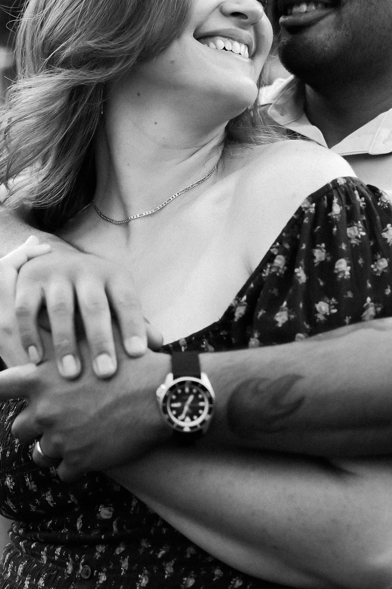 A black and white photo of a smiling couple in close embrace, showing part of their faces and arms. The woman has shoulder-length hair, is wearing a patterned dress, a necklace, and a watch on her wrist. Their hands are clasped together in the foregr