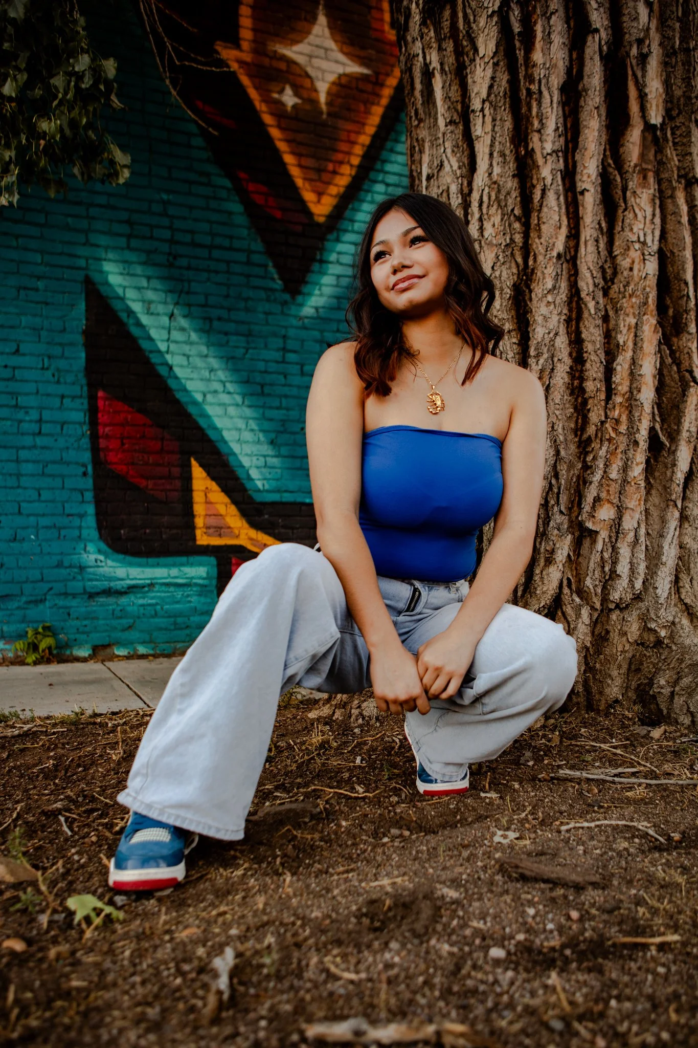 A young woman with shoulder-length dark hair, wearing a blue tube top, light gray baggy jeans, and sneakers, kneeling on the ground near a tree, with a colorful graffiti wall featuring large abstract shapes behind her.