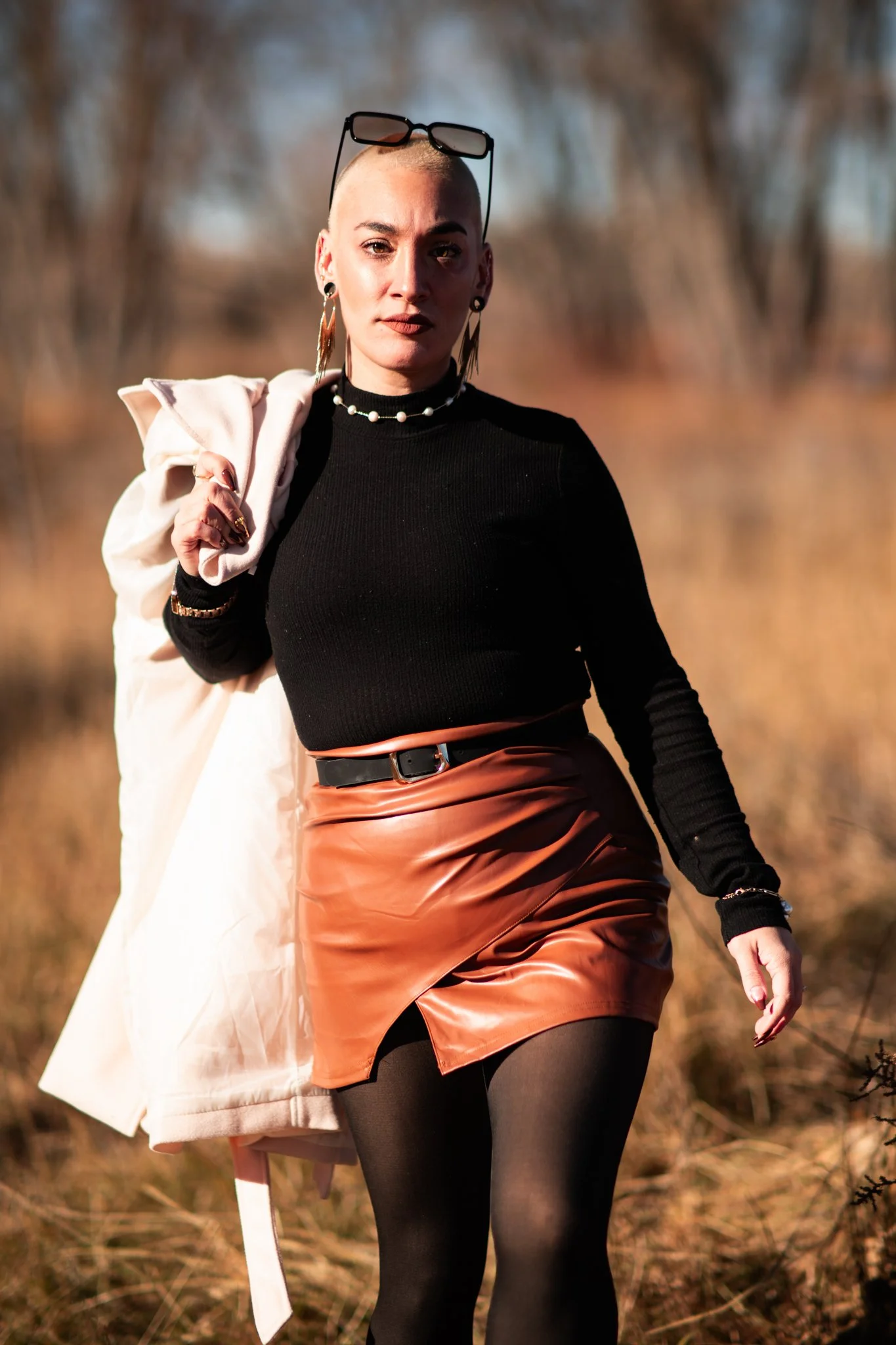 A woman with a shaved head, black turtleneck sweater, leather mini skirt, black tights, and earrings stands outdoors in a natural setting, holding a white jacket over her shoulder, with sunglasses on her head.