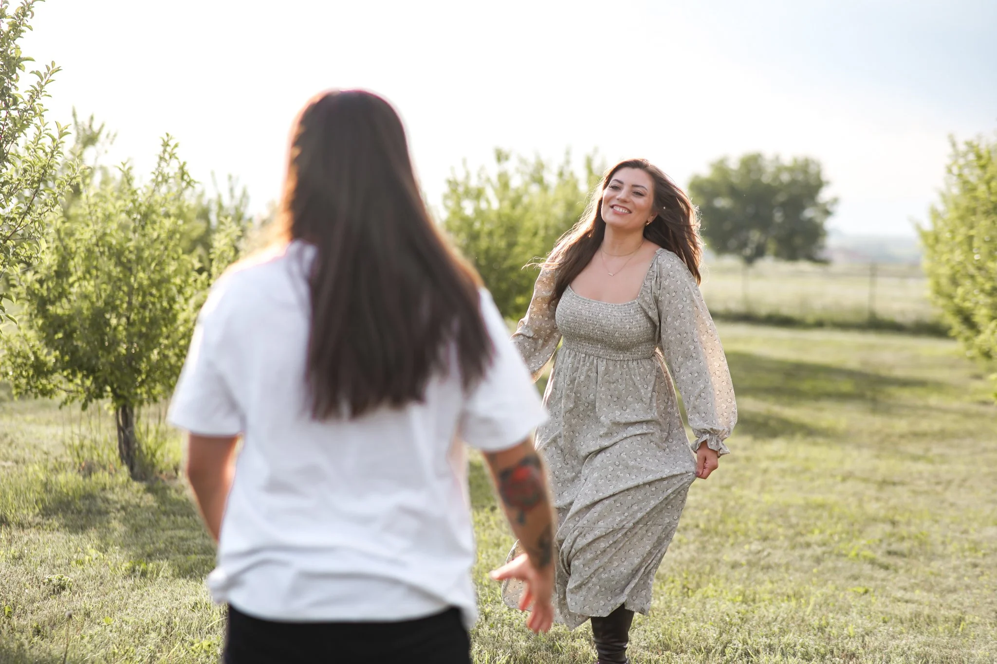 Two women are enjoying a sunny day outdoors among trees, with one woman facing the camera and smiling while the other woman has her back to the camera.