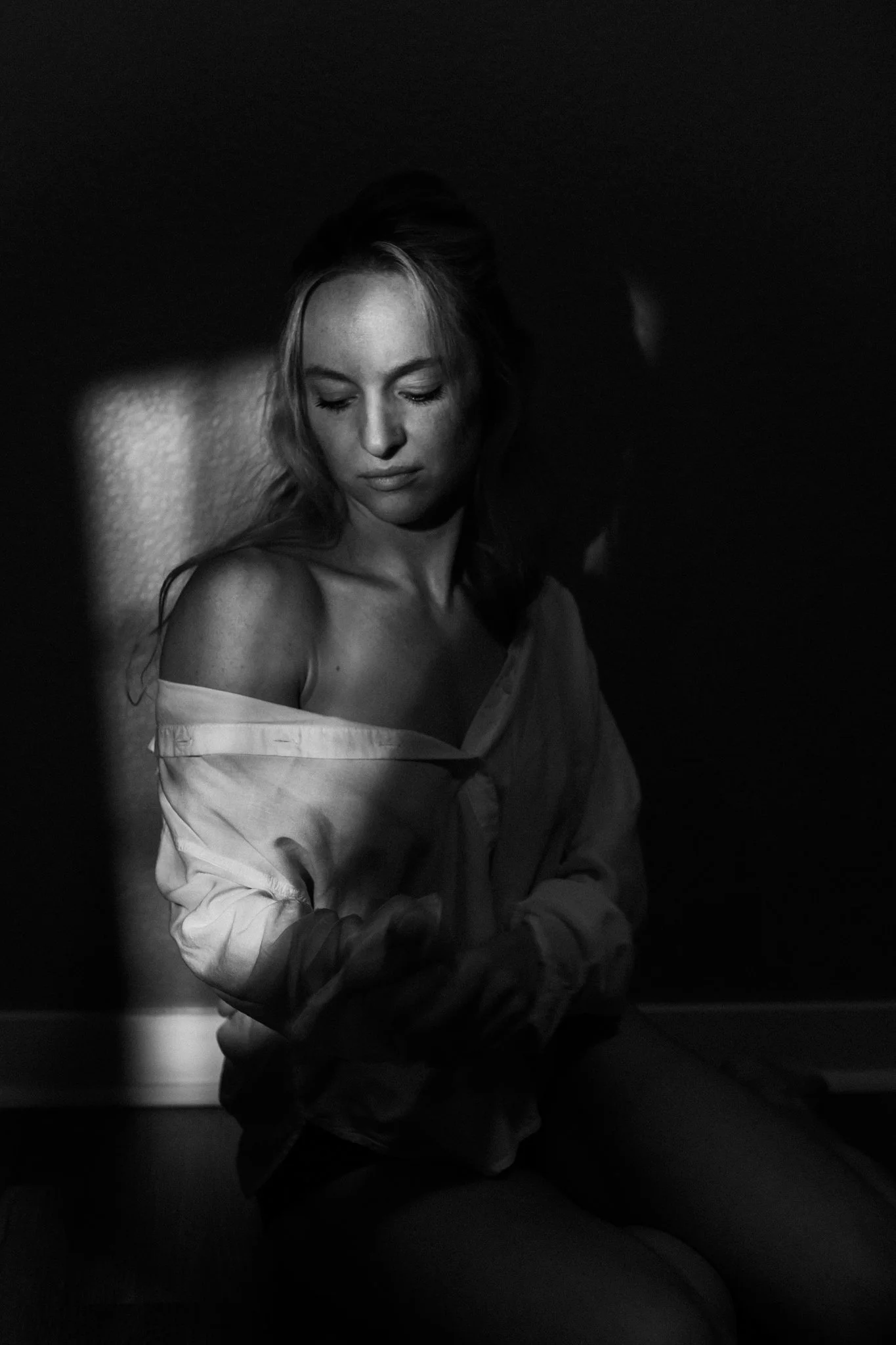 A black-and-white photo of a woman with wavy hair, sitting against a wall with a shadow cast across her face. She is wearing an open shirt with her shoulders exposed, looking down with a calm expression.