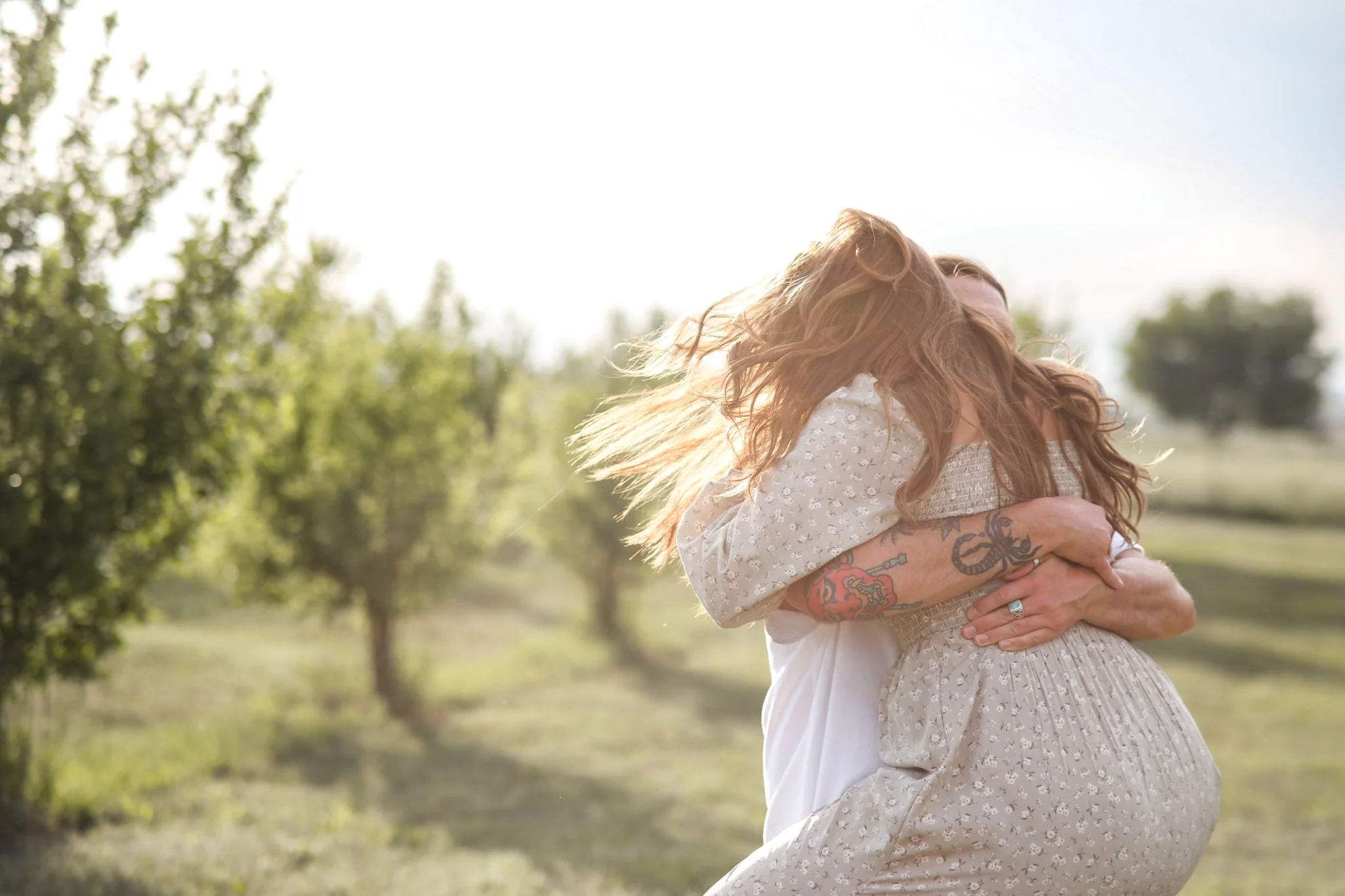 Two people embracing outdoors in a grassy area with trees, sunlight shining in the background.