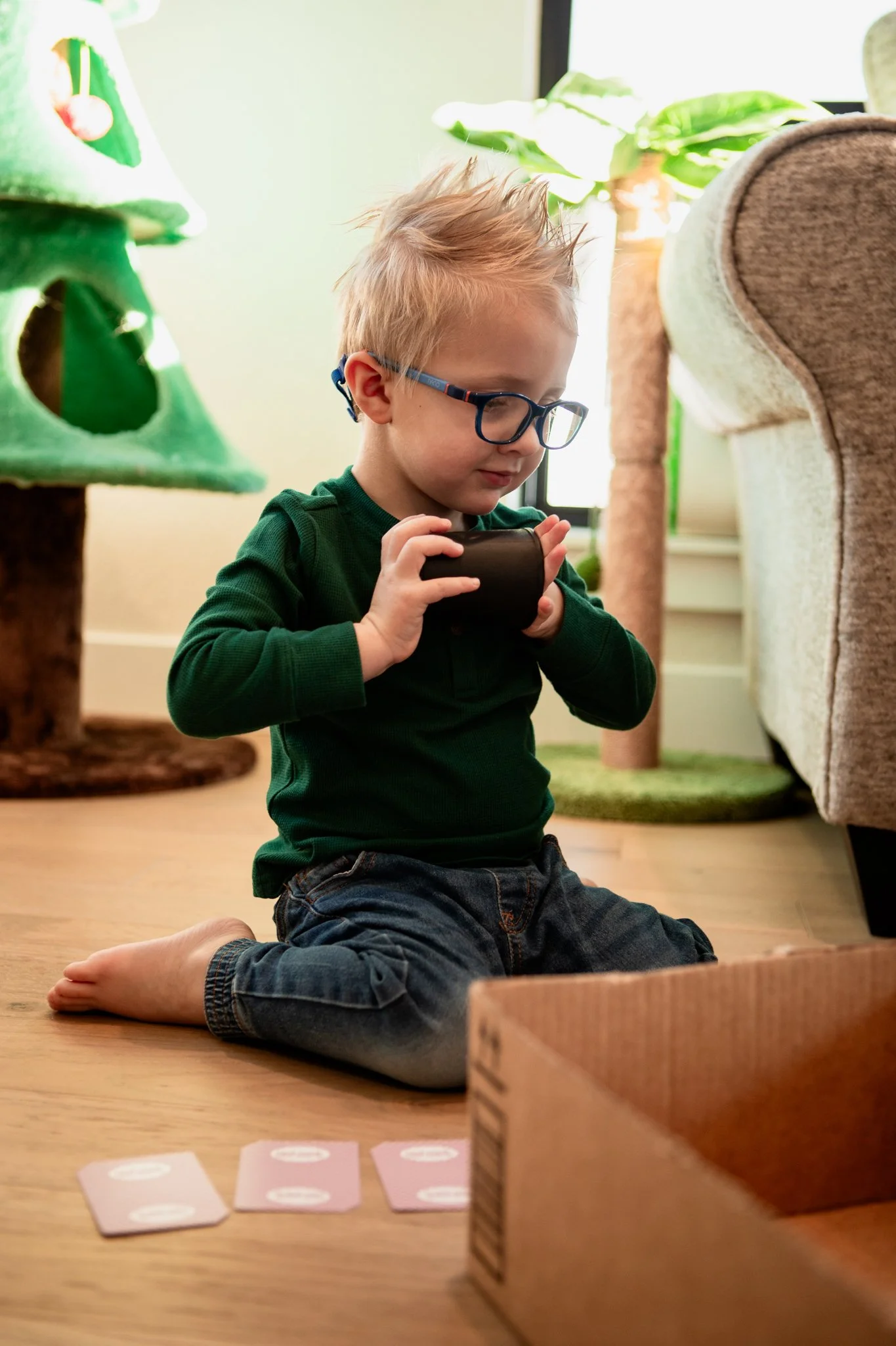 A young boy with glasses, wearing a green shirt and jeans, is sitting on the floor indoors, playing with a black cylindrical object, with four pink cards with white text lying in front of him. There is a cardboard box in the foreground and a decorate