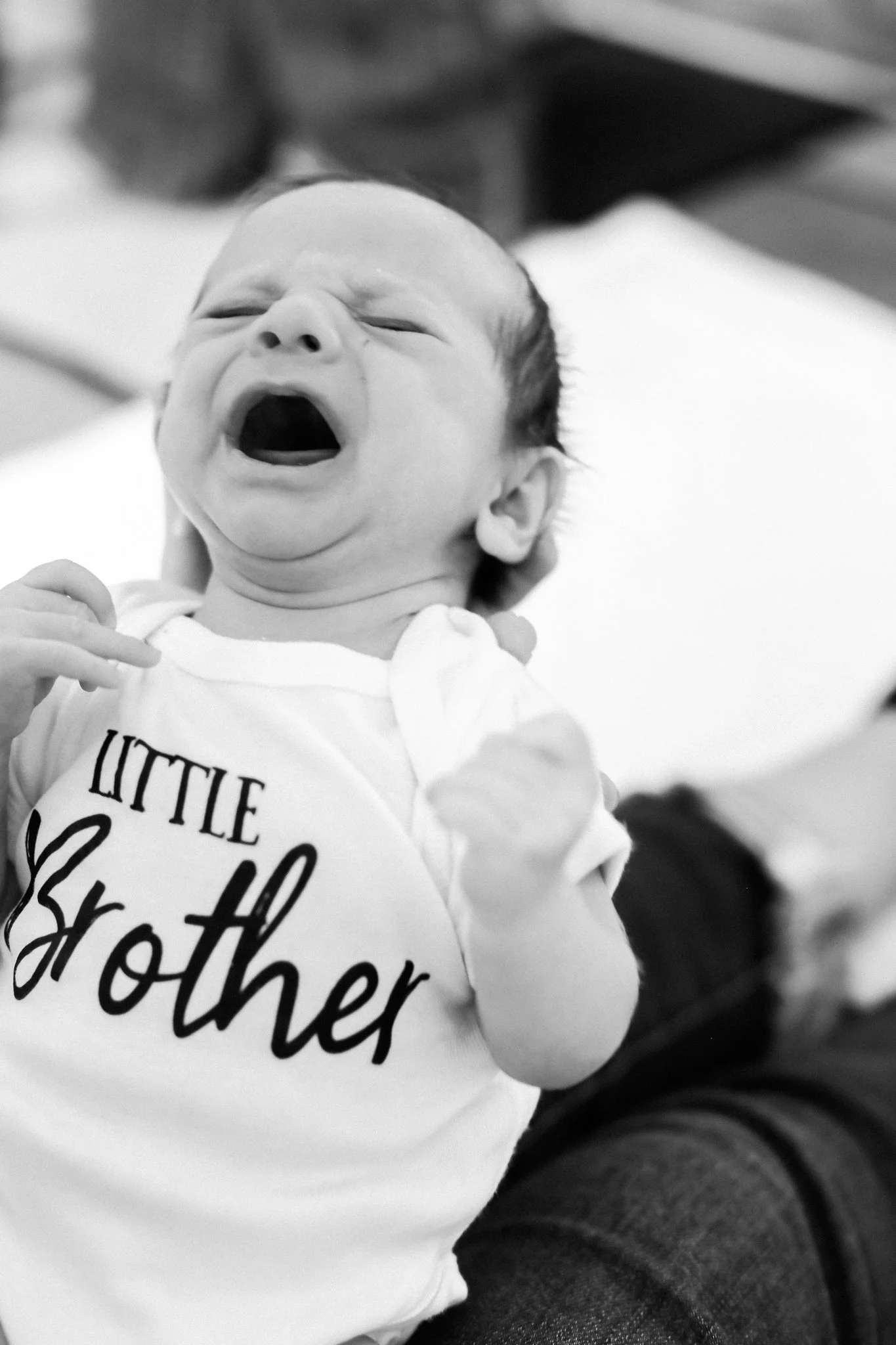 A crying baby wearing a T-shirt that says 'Little Other' on it.