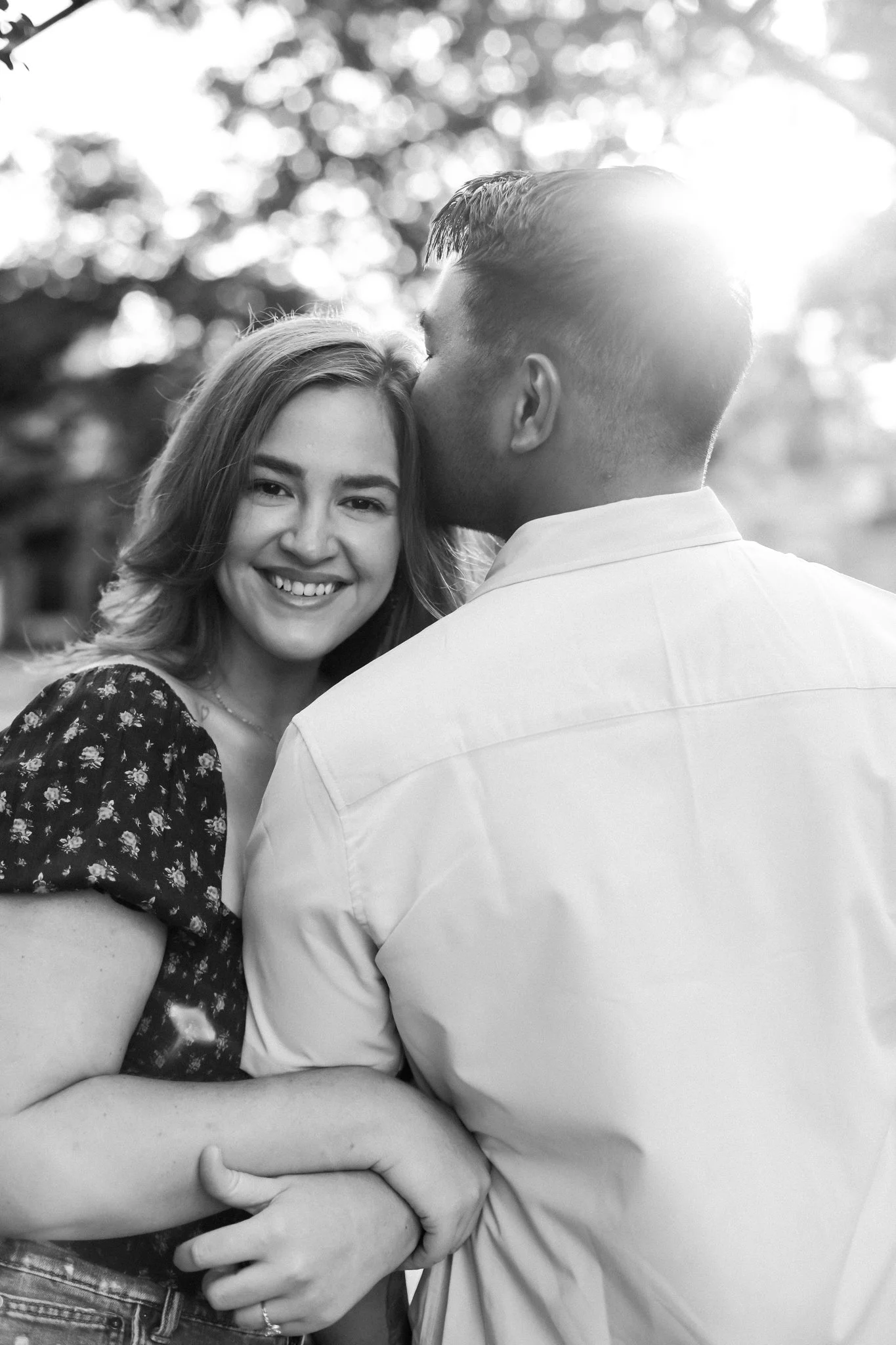 A black and white photo of a smiling woman with long hair, being kissed on the forehead by a man with short hair, outdoors with sunlight and trees in the background.