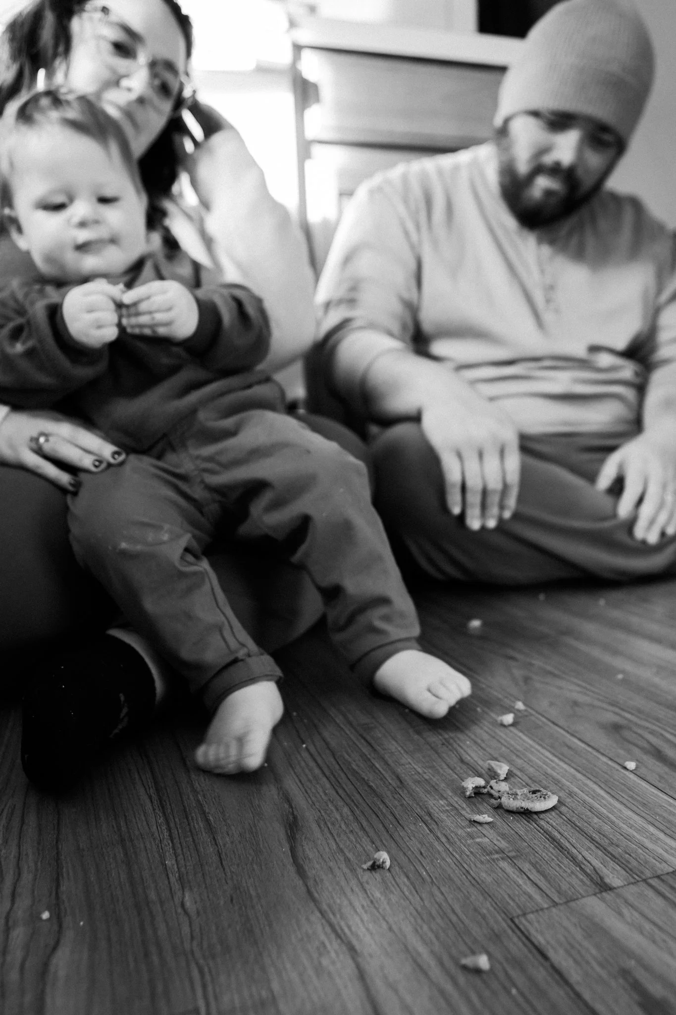 A black-and-white photo of a family sitting on a wooden floor, with crumbs and bitten cookies in front of them. The child is sitting on a woman’s lap, and the man is sitting beside them, looking down.