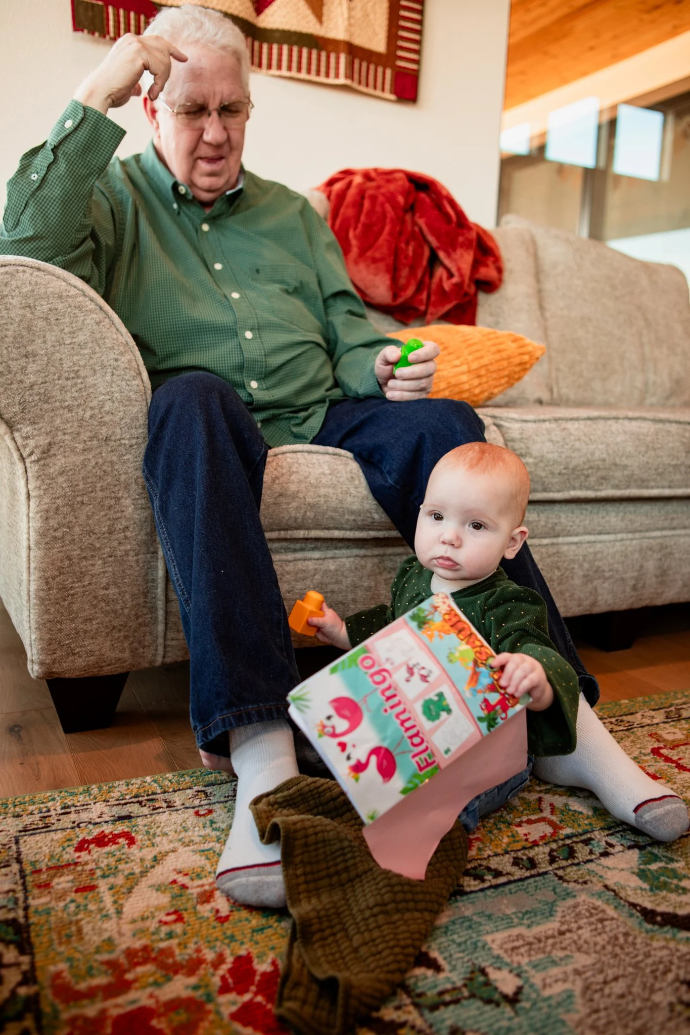 An elderly man with glasses and white hair sitting on a beige couch, frowning and scratching his head while holding green toy. A toddler with red hair, wearing a green dress, sitting on the floor with a Christmas stocking and holding a holiday-themed