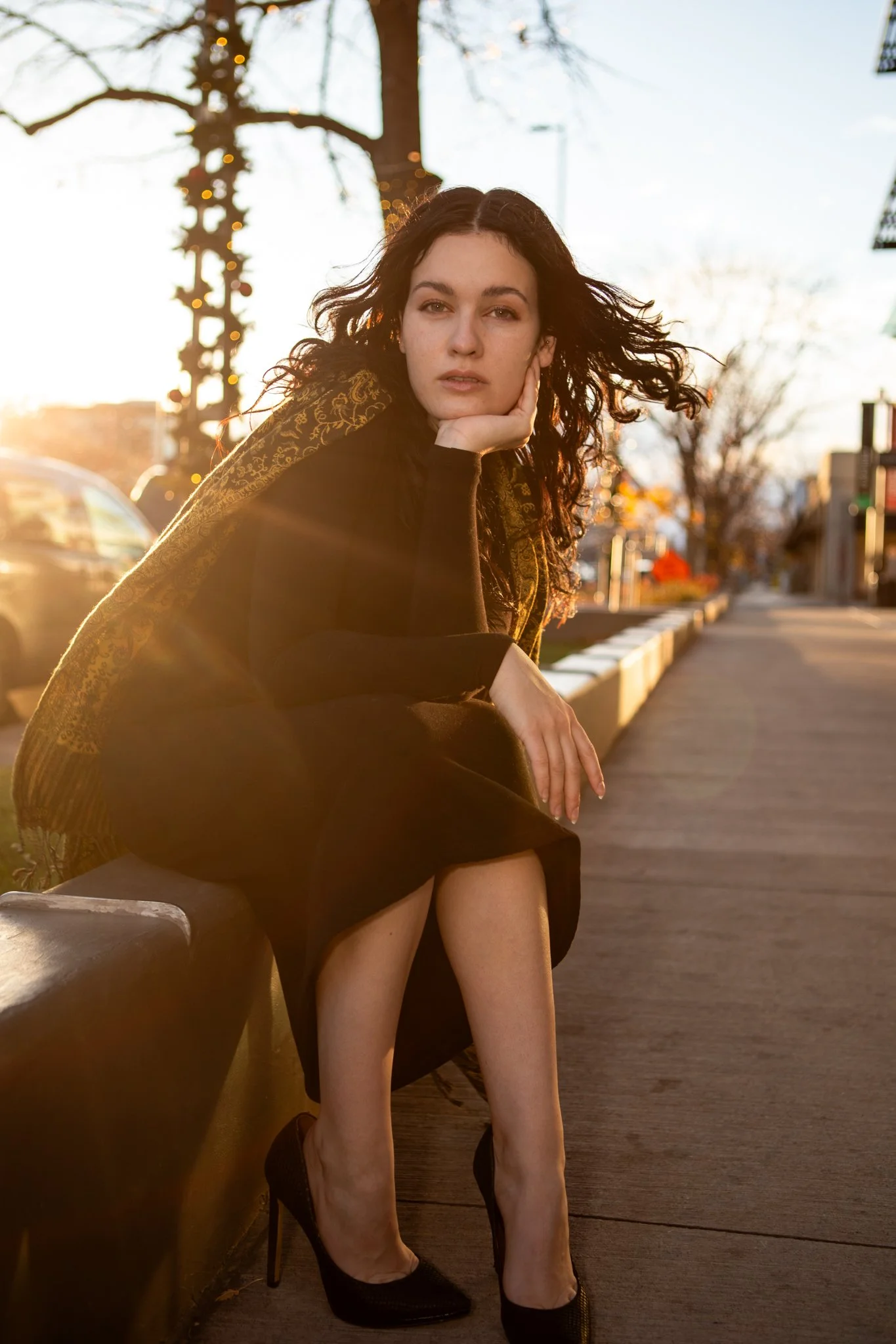 A woman with dark curly hair sitting on a park bench during sunset, wearing high heels and a black dress, with a thoughtful expression, and her hand resting on her face.