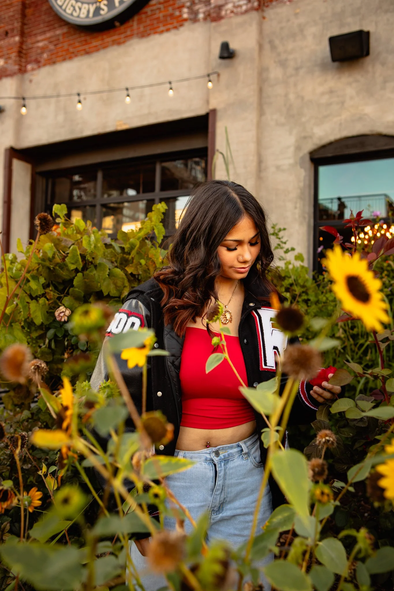 A young woman with dark wavy hair looking down at her phone amidst sunflowers and greenery, with a brick and concrete building in the background.