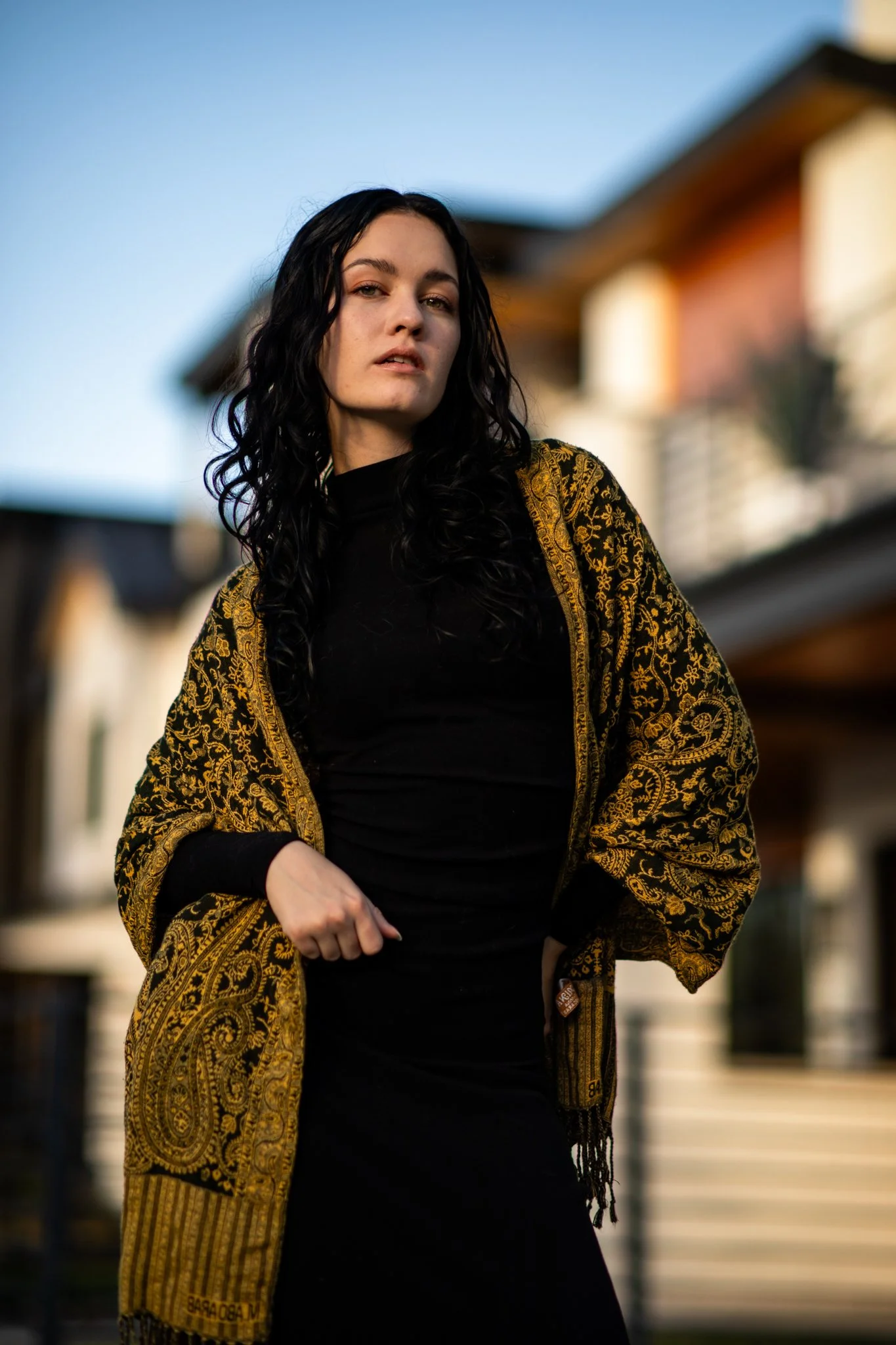 A woman with long black curly hair wearing a black dress and a gold-patterned shawl standing outdoors with a blurred residential background.