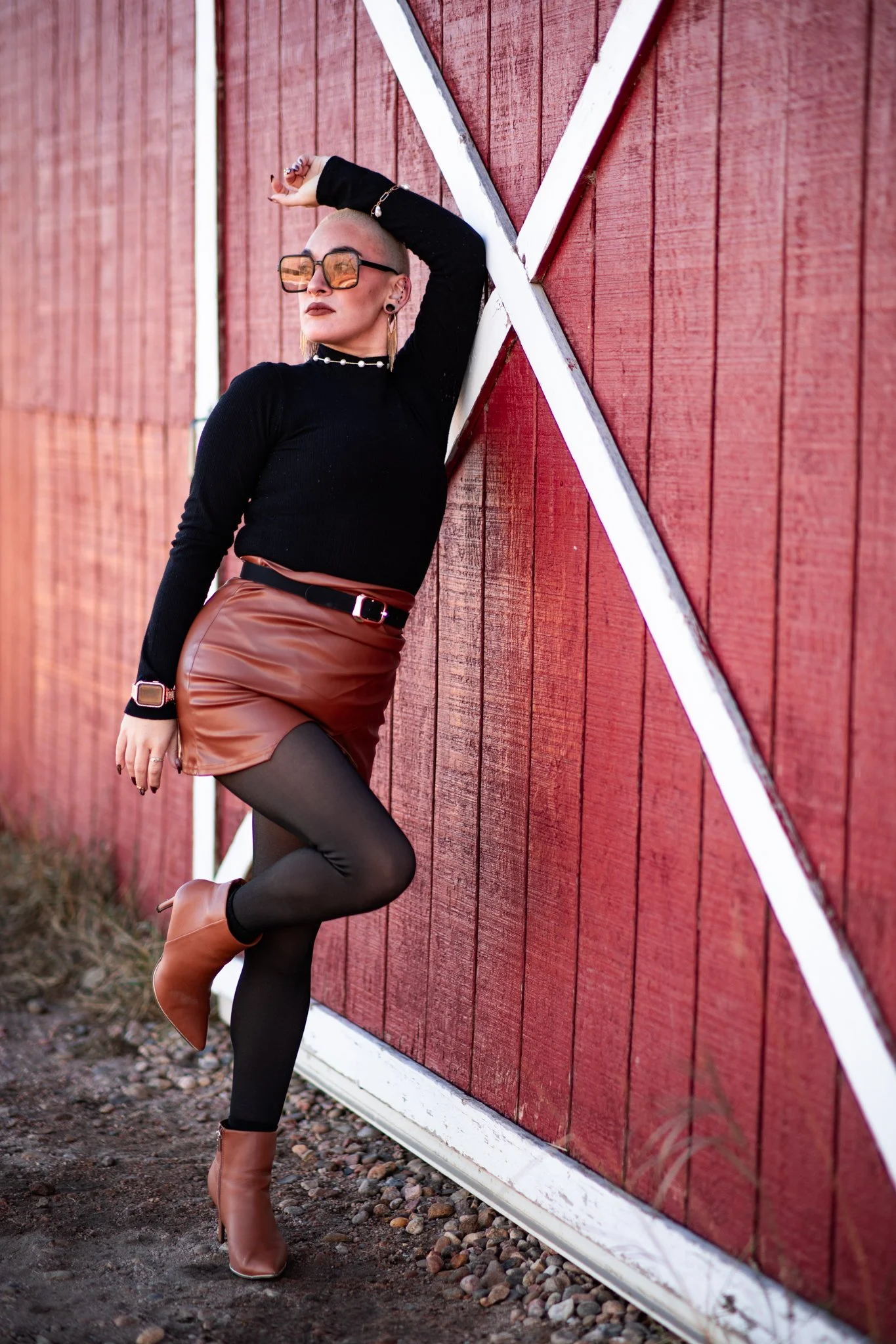 A woman with blonde hair and sunglasses leaning against a red barn wall, dressed in a black top, brown leather skirt, black tights, and brown ankle boots.