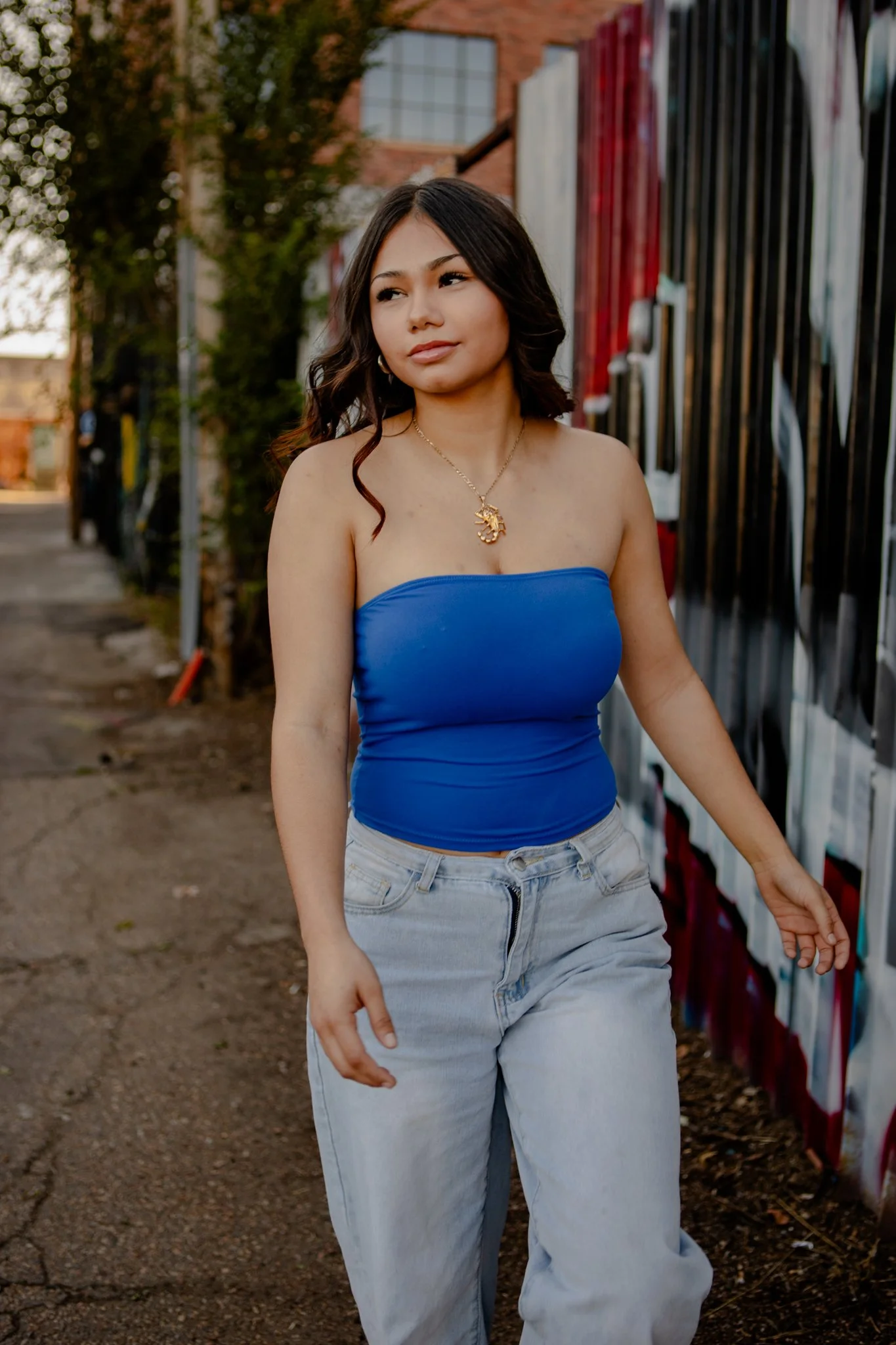 A young woman with dark, wavy hair is walking outdoors beside a graffiti-covered wall. She is wearing a strapless blue top, light-wash jeans, and a gold necklace with a lion head pendant. She has a confident expression and is slightly looking to her 