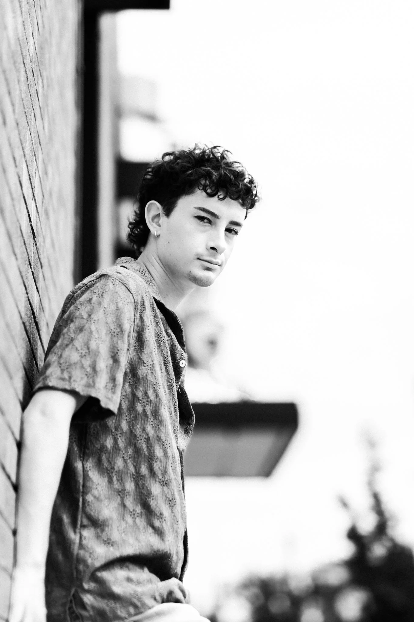 A young man with curly hair and earrings leaning against a brick wall outdoors, looking at the camera with a neutral expression in black and white.