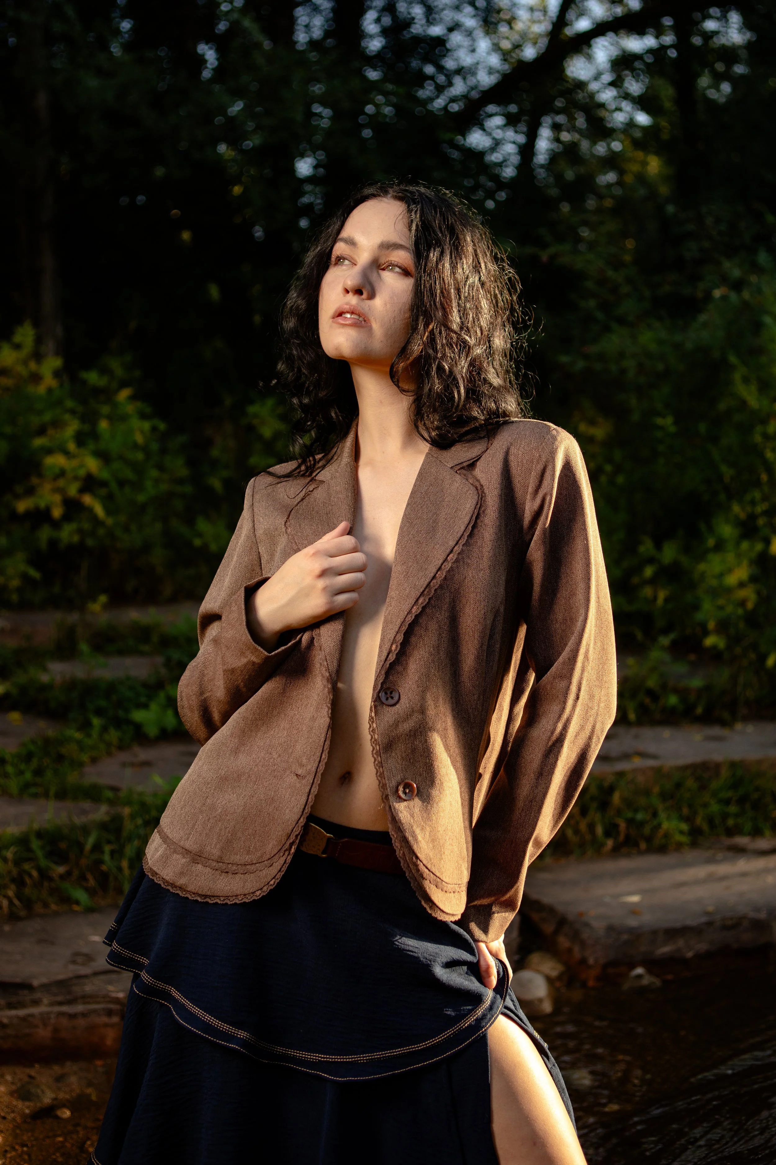 A woman with curly dark hair and light skin, wearing a brown blazer, stands outdoors in a wooded area with sunlight filtering through the trees.