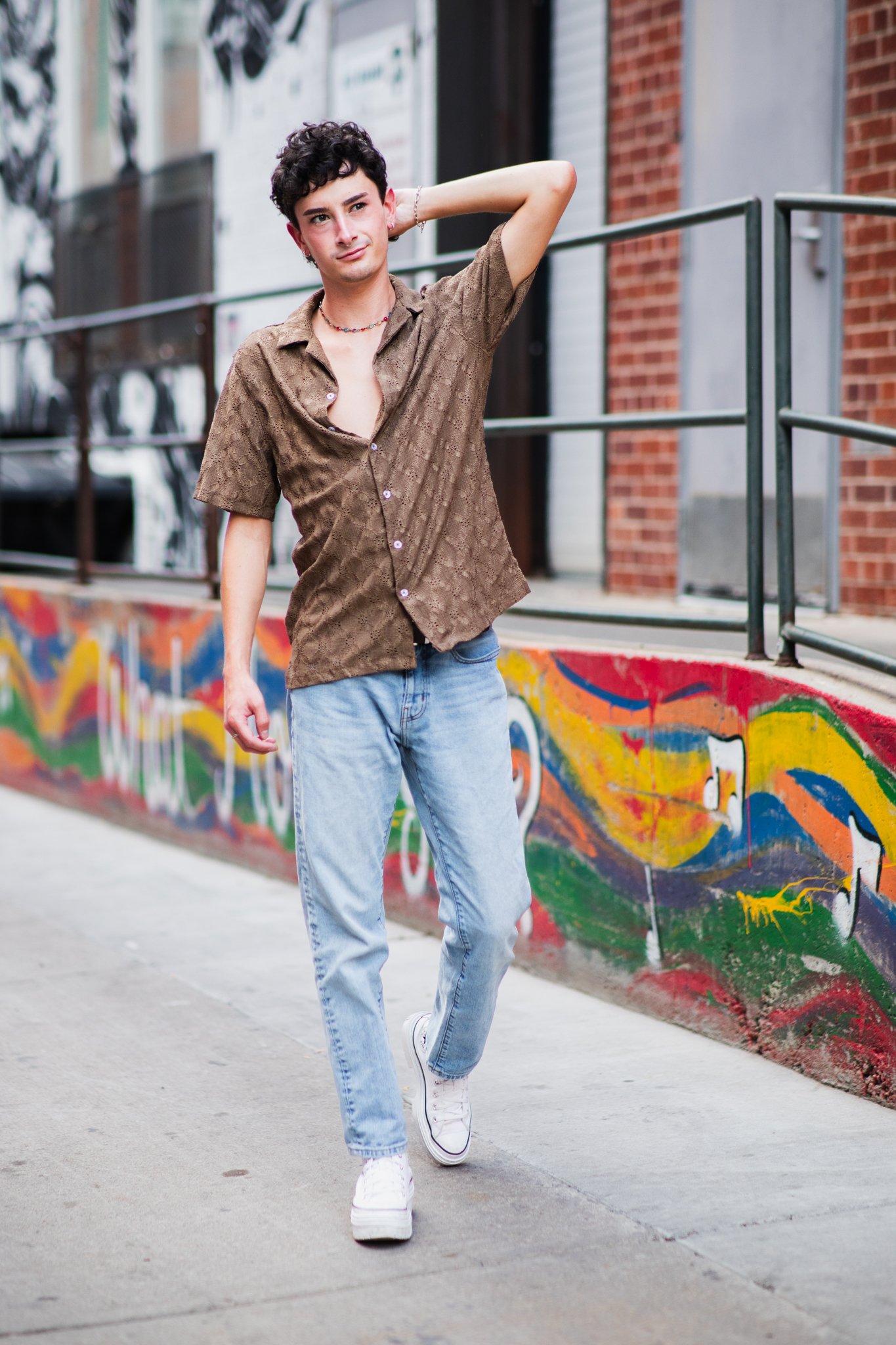 A young person with dark curly hair, wearing a brown patterned short-sleeve shirt, light blue jeans, and white sneakers, walking along a city sidewalk with colorful graffiti on the wall behind them.