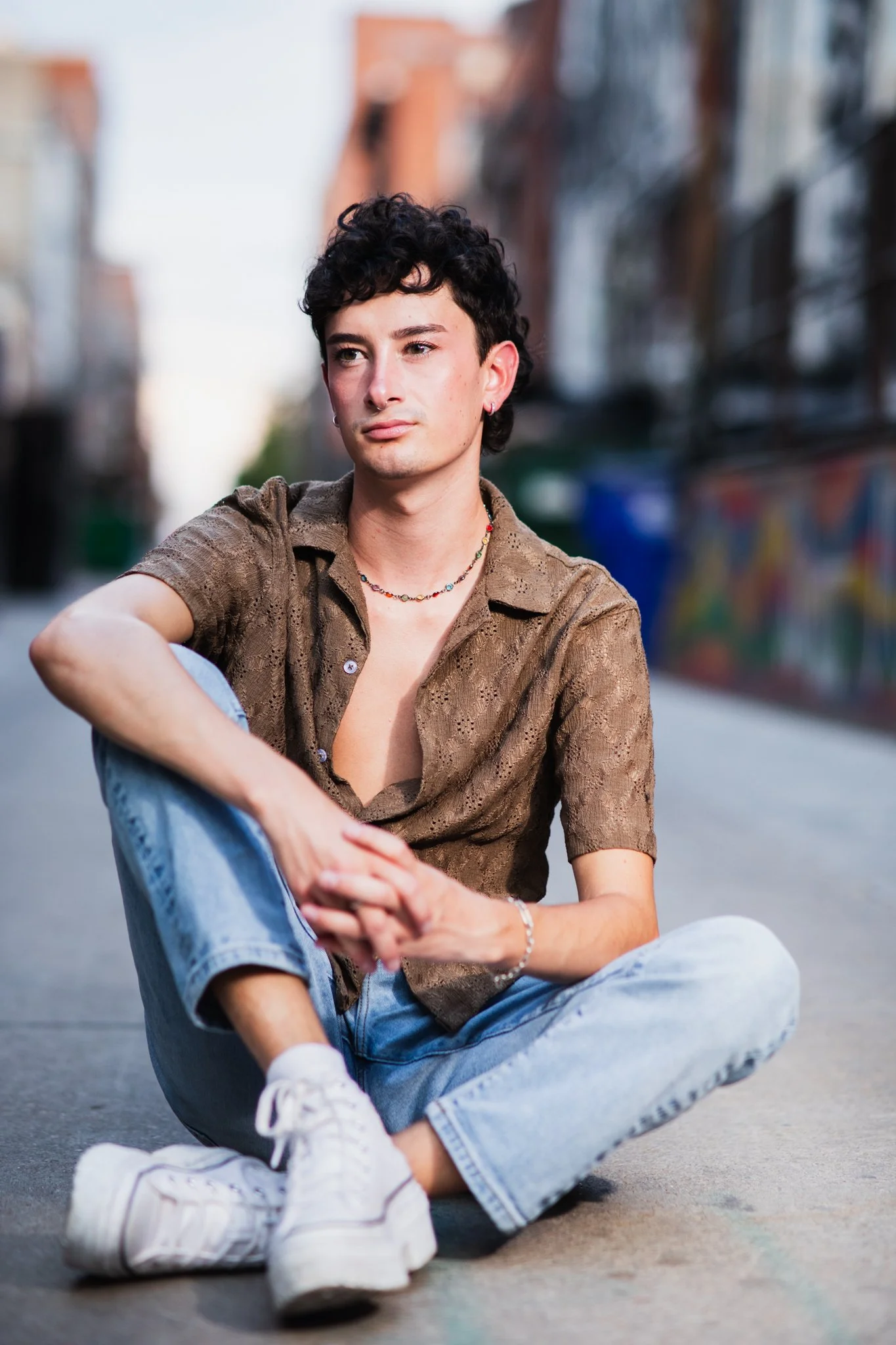 Person sitting on the street with crossed legs, wearing a brown shirt, light blue jeans, and white sneakers, in an urban environment with graffiti in the background.