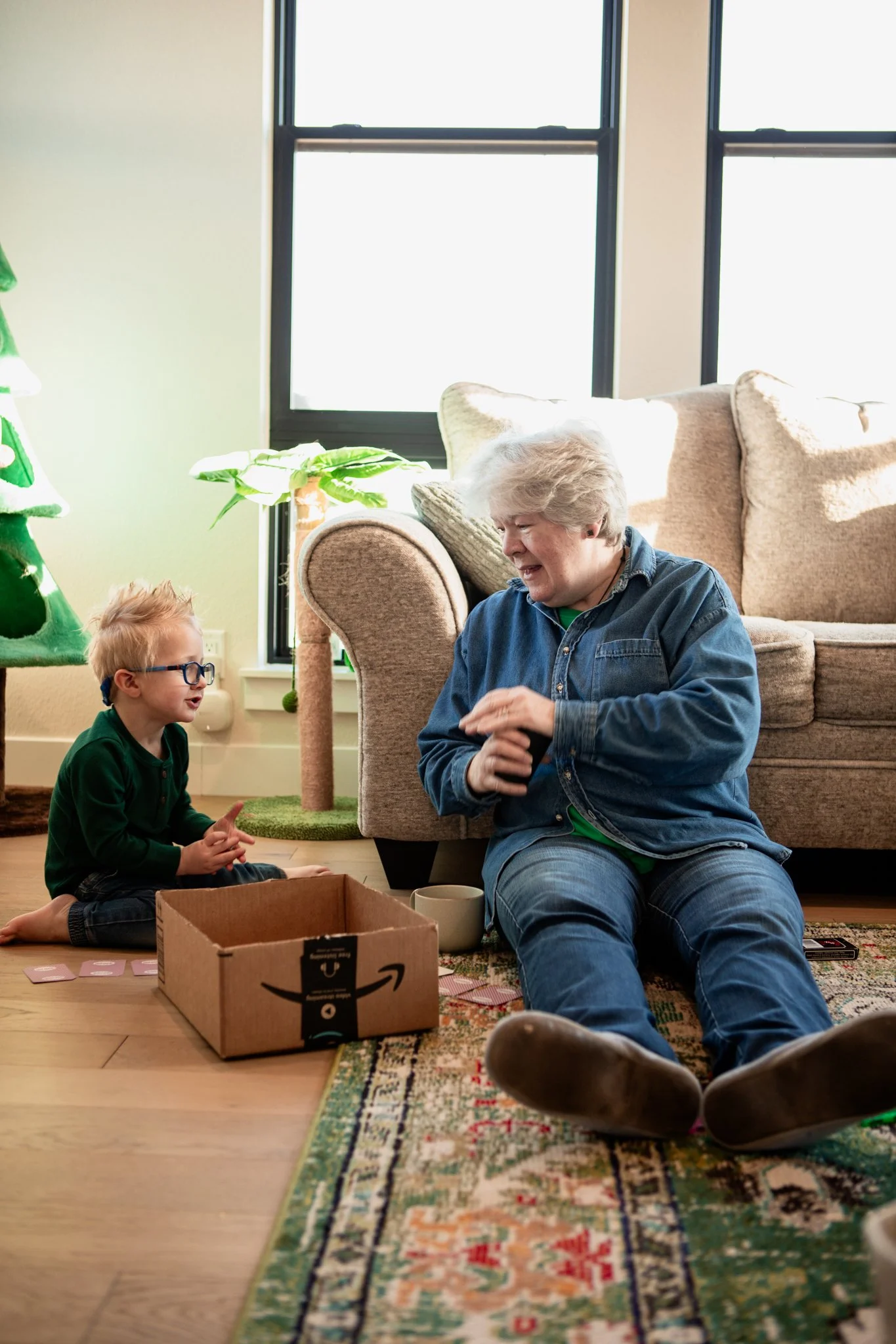 A young boy and an elderly woman sitting on the floor, playing a card game next to a cardboard box and coffee mug in a cozy living room with a Christmas tree.