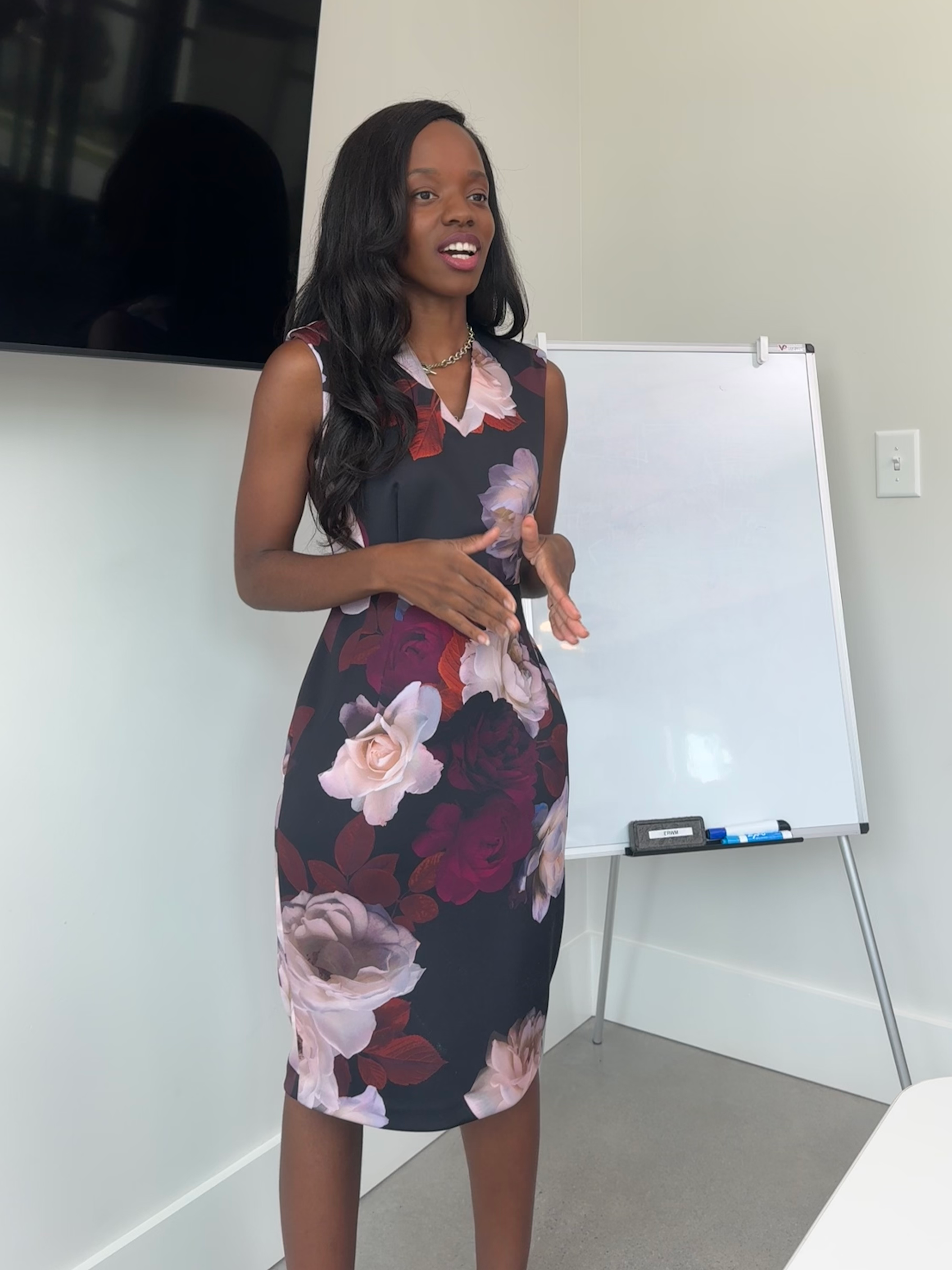 A woman with long dark hair is standing and speaking in front of a whiteboard in a conference room, wearing a sleeveless floral dress.