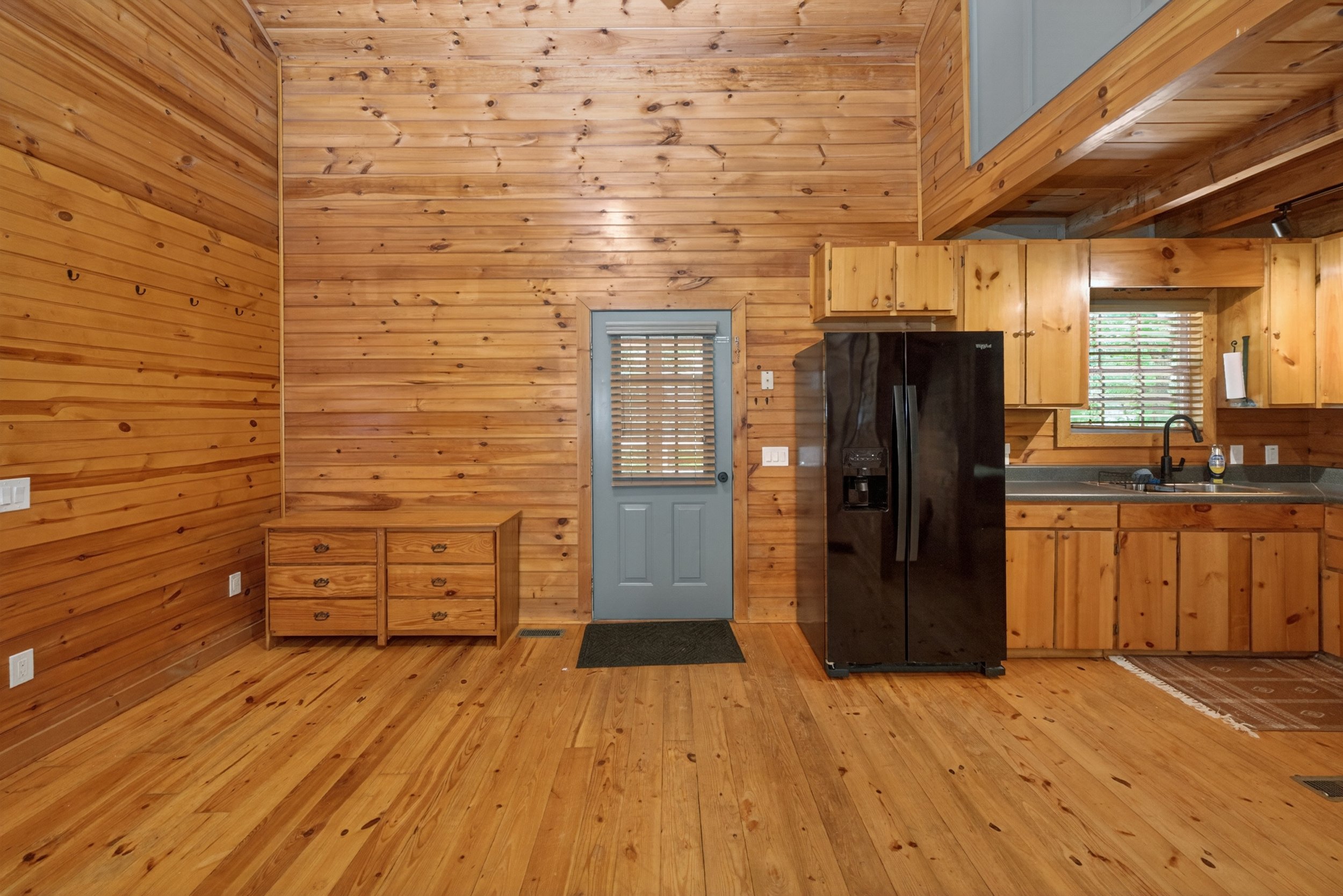 A wooden interior space featuring wood paneling on the walls and floors, a black refrigerator, a window with blinds, a door, a small dresser with multiple drawers, hooks on the wall, and a kitchen sink with a faucet and paper towel holder.
